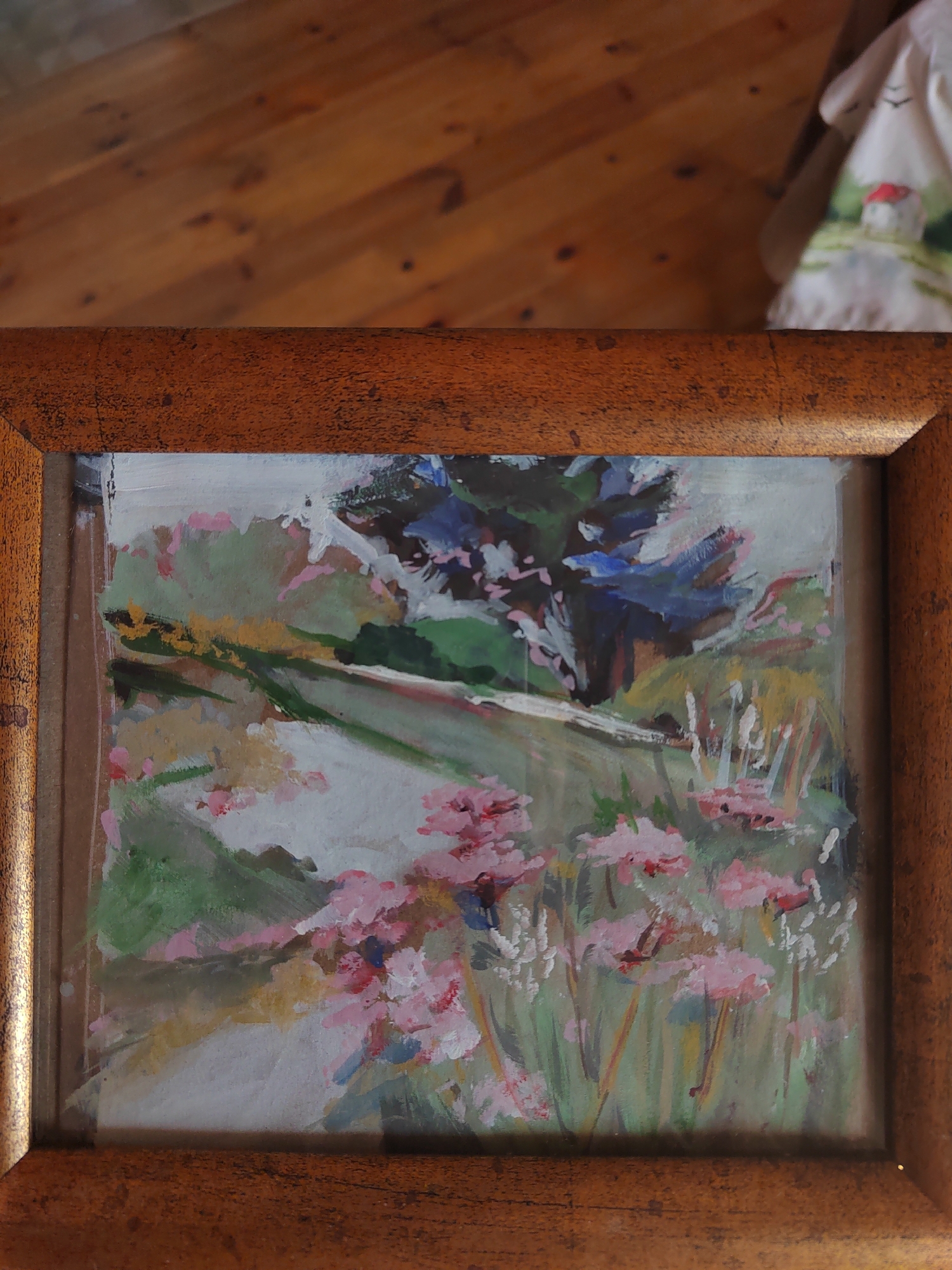 The image shows a framed painting of a floral landscape. The painting is abstract and impressionistic, with bold brushstrokes of pink, green, yellow, and blue. The foreground is filled with clusters of pink flowers, while the background features lush green foliage and a dark blue tree. The painting is set against a wooden floor, and a piece of fabric with a depiction of a small house with a red roof is visible in the upper right corner. The location is not specified within the image or any accompanying metadata.