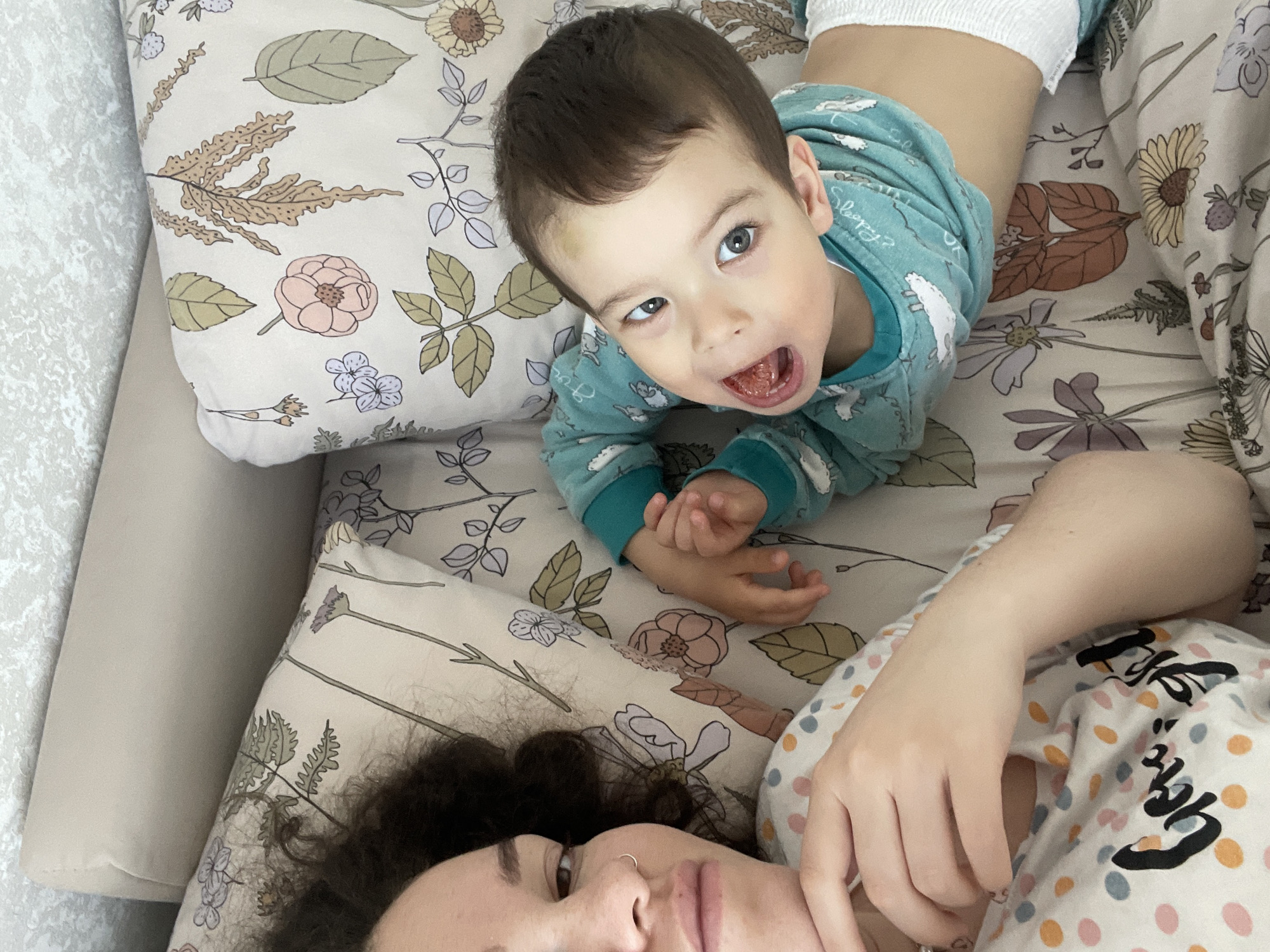 This high-angle image captures an intimate moment between an adult woman and a young child, likely in a home setting. The scene appears to be taking place on a bed or sofa, covered with a light-colored duvet featuring a delicate floral and leaf pattern in muted tones of pink, purple, yellow, and green. A neutral-toned cushion or armrest is visible along the left side.
In the lower half of the frame, an adult woman lies on her back, with her face and upper chest visible. She has dark, curly hair and a subtle nose piercing. Her gaze is directed towards the camera, and her expression is neutral. She wears a light-colored top with colorful polka dots and some dark, stylized, unreadable text.
Positioned above the woman, a young child lies on their stomach, with their head turned towards the camera. The child has dark brown hair, bright blue eyes, and an open-mouthed, wide-eyed expression that suggests surprise or playfulness. They are wearing a teal long-sleeved top with white patterns, and the text "Sleepy? Ok!" is partially visible on the fabric. A white diaper or pull-up is visible around their waist.
The lighting indicates daytime, contributing to the bright and cozy atmosphere of the indoor setting. The image conveys a sense of connection and warmth between the two subjects, captured in a casual, everyday moment.