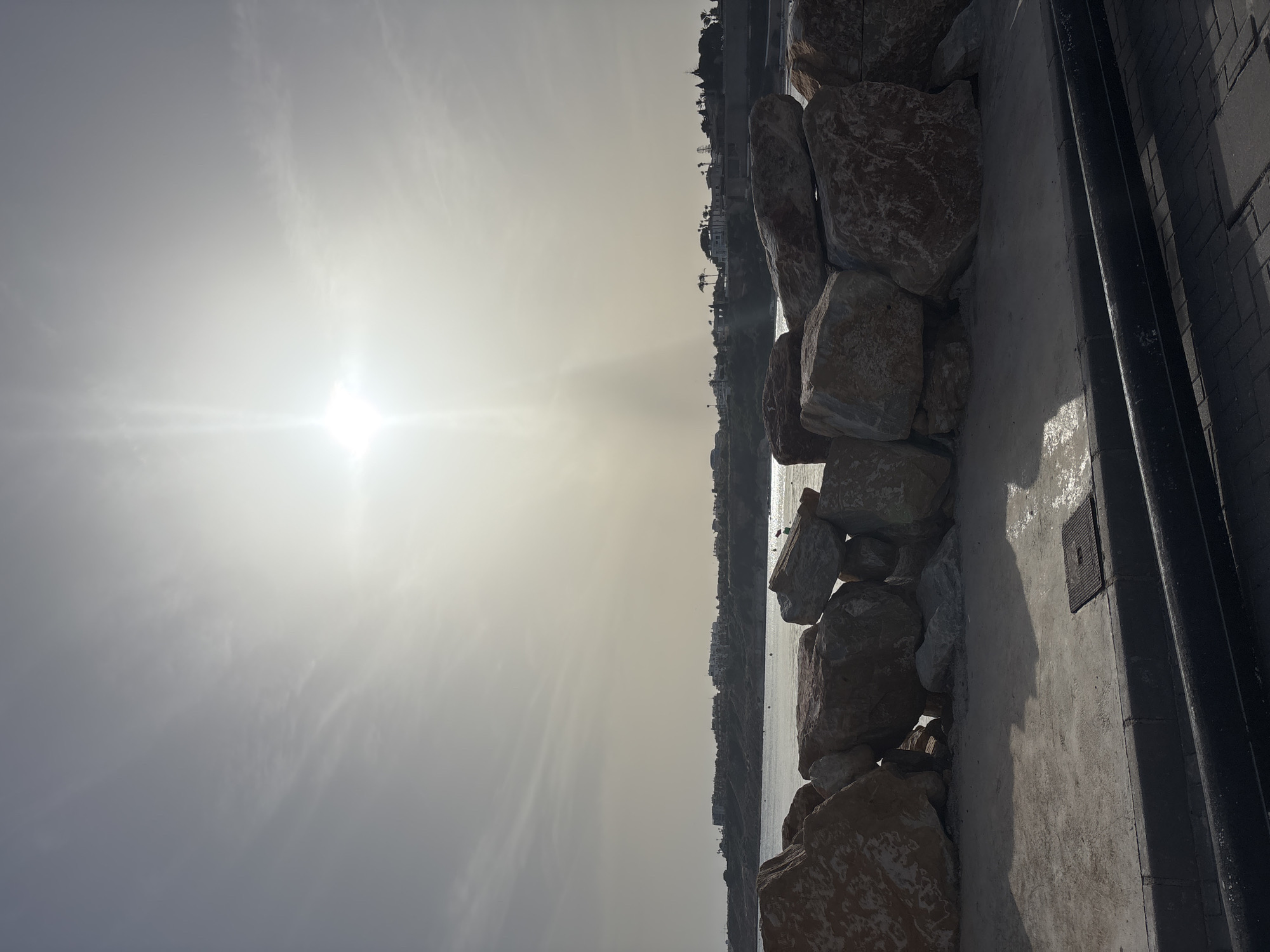 The image depicts a bright, sunny day at a coastal location, likely Urbanización Cabo Roig, Spain. In the foreground, a rough stone wall made of large, irregular rocks forms a barrier. Beyond the wall, a calm body of water, presumably the sea, stretches towards a distant shoreline. Along the shoreline, a cluster of buildings, likely residential properties, are visible against a hazy sky. The sun is high in the sky, casting a bright light and creating a lens flare effect. The overall scene conveys a serene coastal atmosphere.