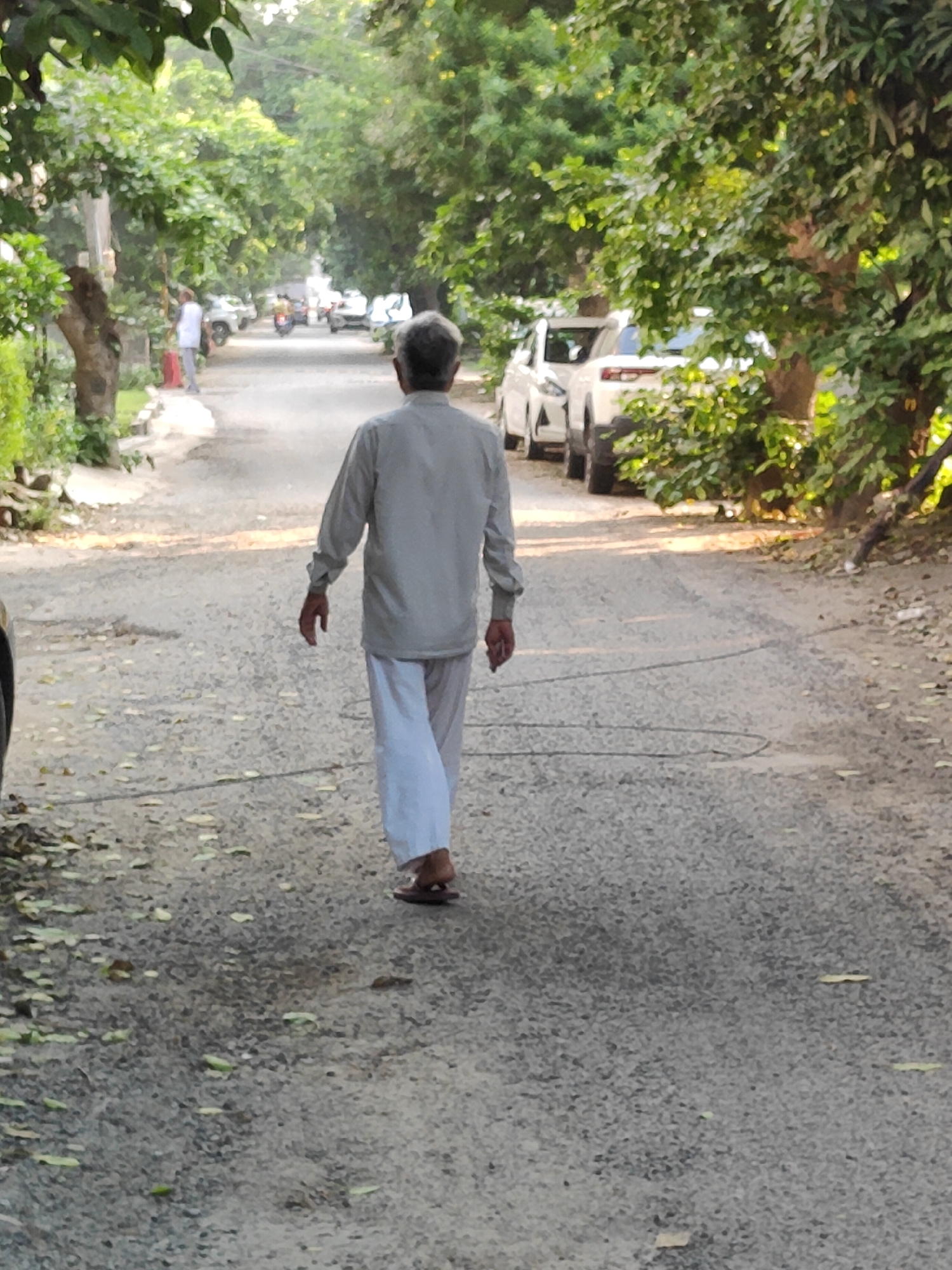 An elderly man with grey hair is walking away from the camera down a paved road lined with trees. He is wearing a light-colored, long-sleeved shirt and white pants. He has brown sandals on his feet. The road is slightly uneven and has scattered leaves on it. In the background, there are parked cars and a few other people walking. The scene appears to be in a residential area with dense foliage. The lighting suggests it is daytime. This location is likely Gurgaon, India, given the common attire and the style of vehicles visible.