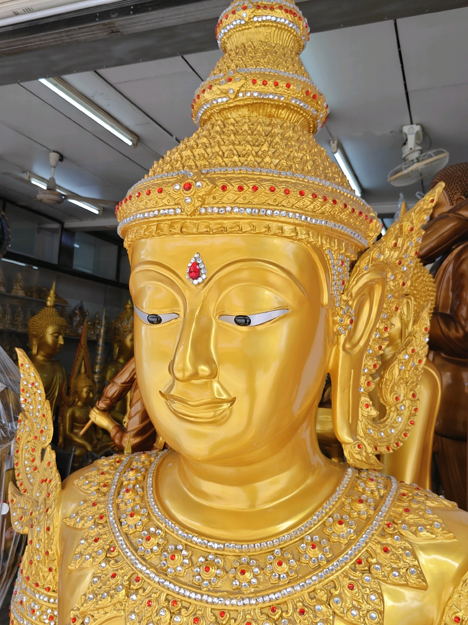 The image shows a close-up of a large, golden statue of a Buddha. The statue is elaborately decorated with red and clear gemstones, particularly around the headdress and collar. The statue's eyes are open and painted black. Behind the main statue, other golden Buddha statues are visible in the background, as well as fluorescent lights and ceiling fans on the ceiling. The overall impression is of a religious or cultural setting, likely a temple or a shop selling religious artifacts. The style of the statue suggests it is from Southeast Asia, possibly Thailand, given the request to include Bangkok.