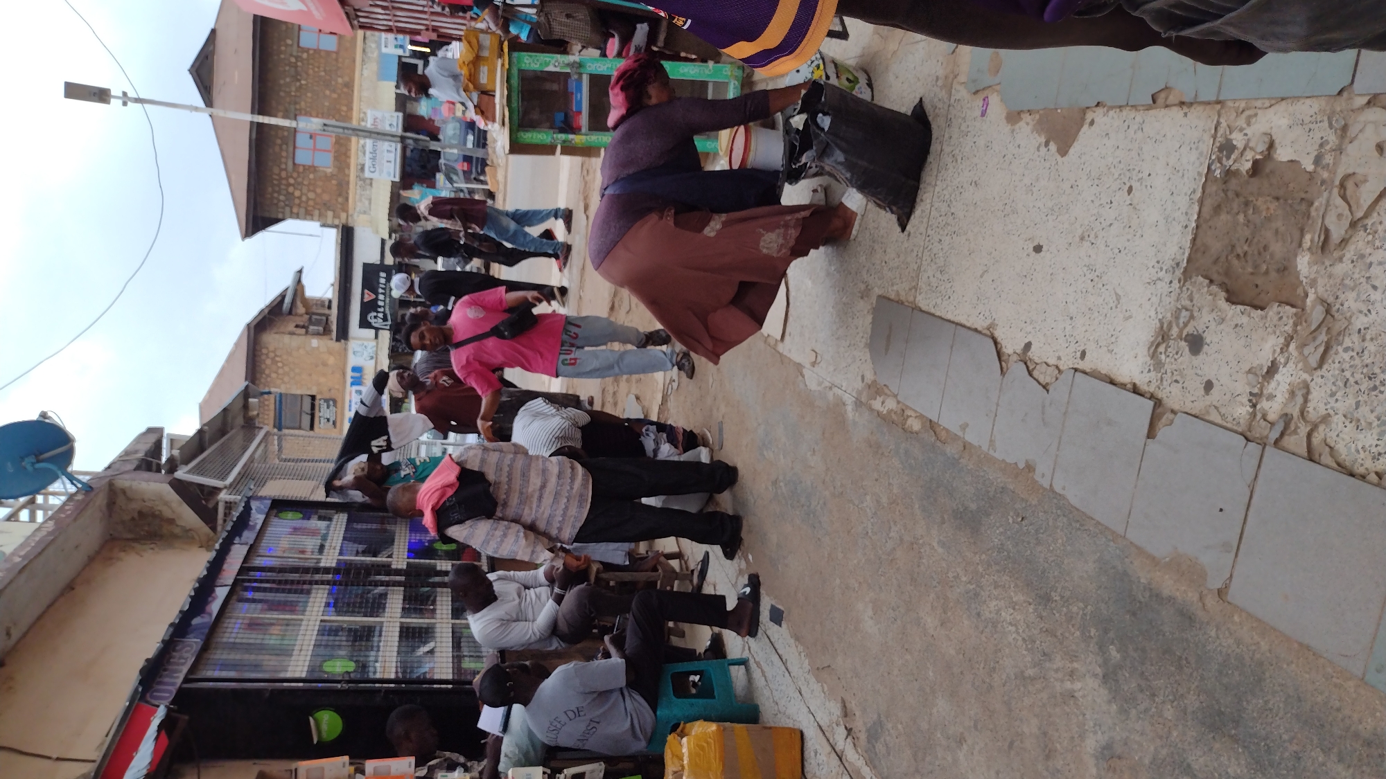 The image captures a street scene in Jos, Nigeria. A group of men are sitting on stools along the edge of a storefront with glass windows, some looking at their phones. A woman with a red headscarf, wearing a purple shirt and brown skirt, carries a black bag as she walks down the sidewalk. Other people are walking in the background. Buildings are visible on either side of the street, with a light pole visible on the left. The sidewalk is damaged, and the ground is worn.
