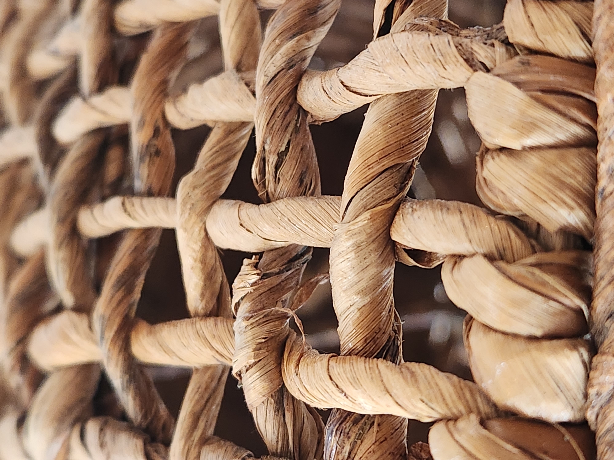 This media file is a close-up, macro photograph of a woven basket, likely made from natural fibers such as rattan or hyacinth. The texture of the woven material is the primary subject, with thick, twisted strands creating an intricate pattern of interlacing loops and solid sections.
The basket appears to be empty, and no activity or event is taking place. The focus is entirely on the texture and construction of the basket itself. The lighting suggests it might be indoors, with a soft, diffused light illuminating the fibers. There are no visible cues about the time of day, weather, or specific location beyond the general context provided. The colors are predominantly earthy tones of beige, brown, and tan, with variations in shade due to the natural material and the way light plays on the textured surface.
Notable details include the detailed rendering of the twisted fibers, revealing individual strands and the slight wear or imperfections that add to the natural character of the object. The depth of field is shallow, with some parts of the basket in sharp focus and others softly blurred, creating a sense of depth and highlighting the three-dimensional nature of the weave. There is no visible text in the image. The scene is static and focuses solely on the tactile and visual qualities of the woven object.