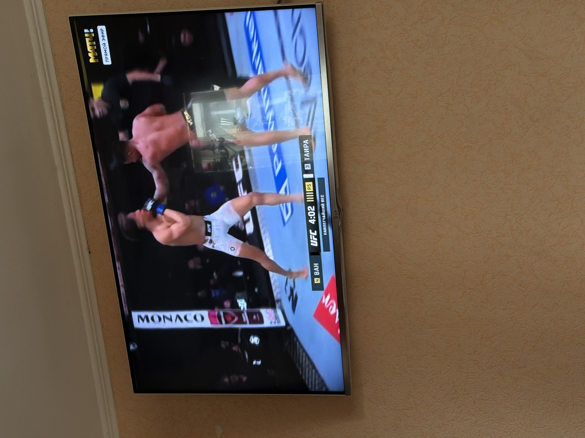The image shows a TV screen displaying a UFC fight. The fight is between two male fighters. One fighter, wearing white shorts, is throwing a punch at the other fighter who is shirtless. The fight is taking place in Monaco. The TV is showing a broadcast from the Russian TV channel "Match!". The lower portion of the screen shows the names of the fighters "BAH" and "ТАИРА". The time on the screen reads 4:02. The TV is mounted on a wall with a beige, textured wallpaper. There is no indication of this image being in Beryozovka, Russia.