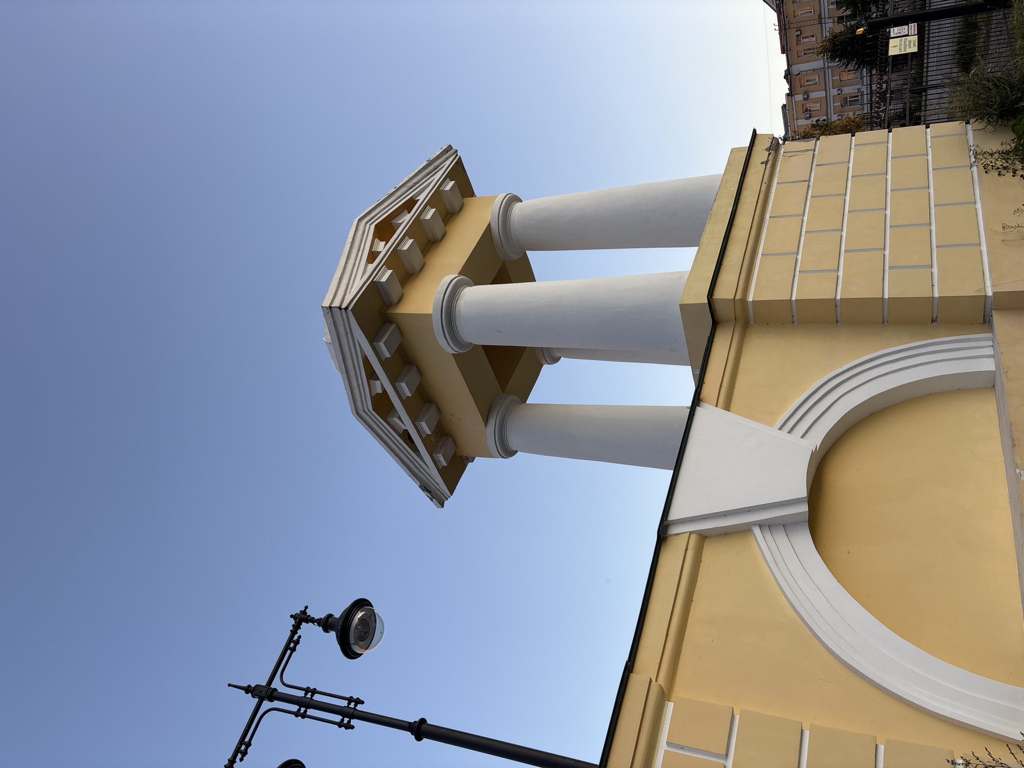 The image shows a low-angle view of a yellow-colored building with classical architectural elements, set against a clear blue sky. The building features a prominent white-capped roof supported by white columns. The facade includes decorative white molding, an arched recess, and a patterned texture resembling bricks. To the left, a black lamppost with an ornate design and a glass-enclosed lamp is visible. In the background, a multi-story building and a black metal fence can be seen. The scene appears to be an outdoor public space, possibly a park or a street in a city. The style of architecture and the presence of a lamppost suggest a historical or culturally significant area. The image quality is clear, with good lighting and focus. There are no people or animals in the frame.
