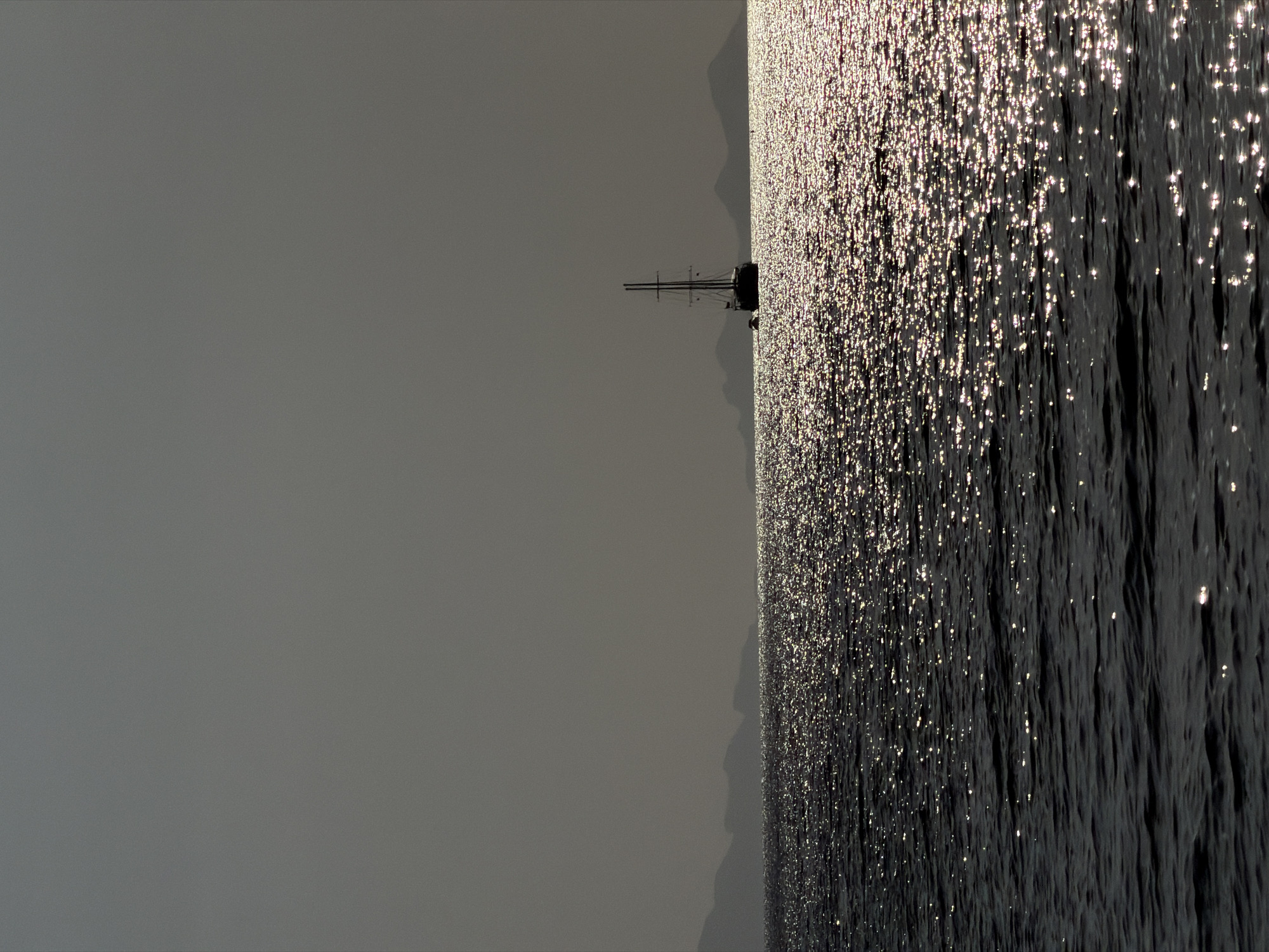 A sailboat is anchored on the calm sea, with the sun reflecting off the water's surface, creating a shimmering effect. In the background, distant islands with silhouetted mountains are visible, partially obscured by a hazy sky. The scene is tranquil, with no people or discernible activity other than the gentle movement of the water and the stationary vessel. The location is the sea off the coast of Ko Samui District, Thailand, likely during either dawn or dusk given the lighting conditions.