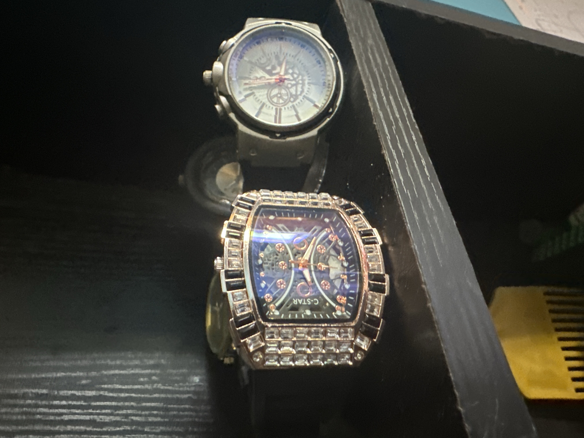 The image shows two watches on a black shelf. The watch on the left is rectangular with a dark blue face, embellished with rhinestones and black stones around the bezel. The watch on the right is round with a silver-toned face and a silver-toned band. The background is dark and out of focus, with some vertical lines visible, suggesting a textured surface. Below the shelf, a portion of a yellow object with vertical ridges is visible, possibly a comb or a similar grooming tool. The image appears to be taken in a domestic setting. There is no information available to link this image to the city of Jauro Yinu, Nigeria.