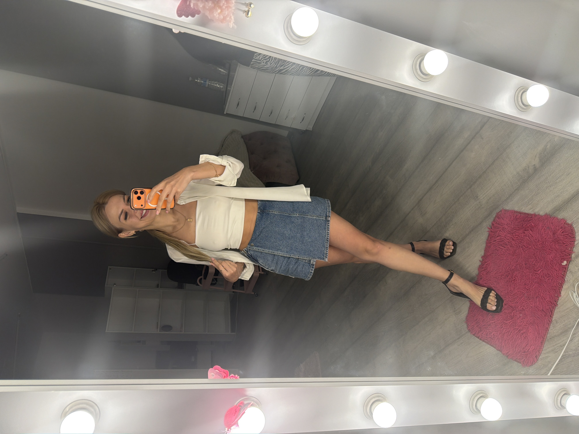 A woman is taking a mirror selfie. She is wearing a white tube top, a white unbuttoned shirt, a denim skirt, and black heels. She has blonde hair pulled back in a ponytail and is holding an orange smartphone. The mirror is part of a vanity with lights. In the reflection, a room is visible with shelves, a dresser, and furniture. The floor is wood-paneled. The location is not identifiable from the image.