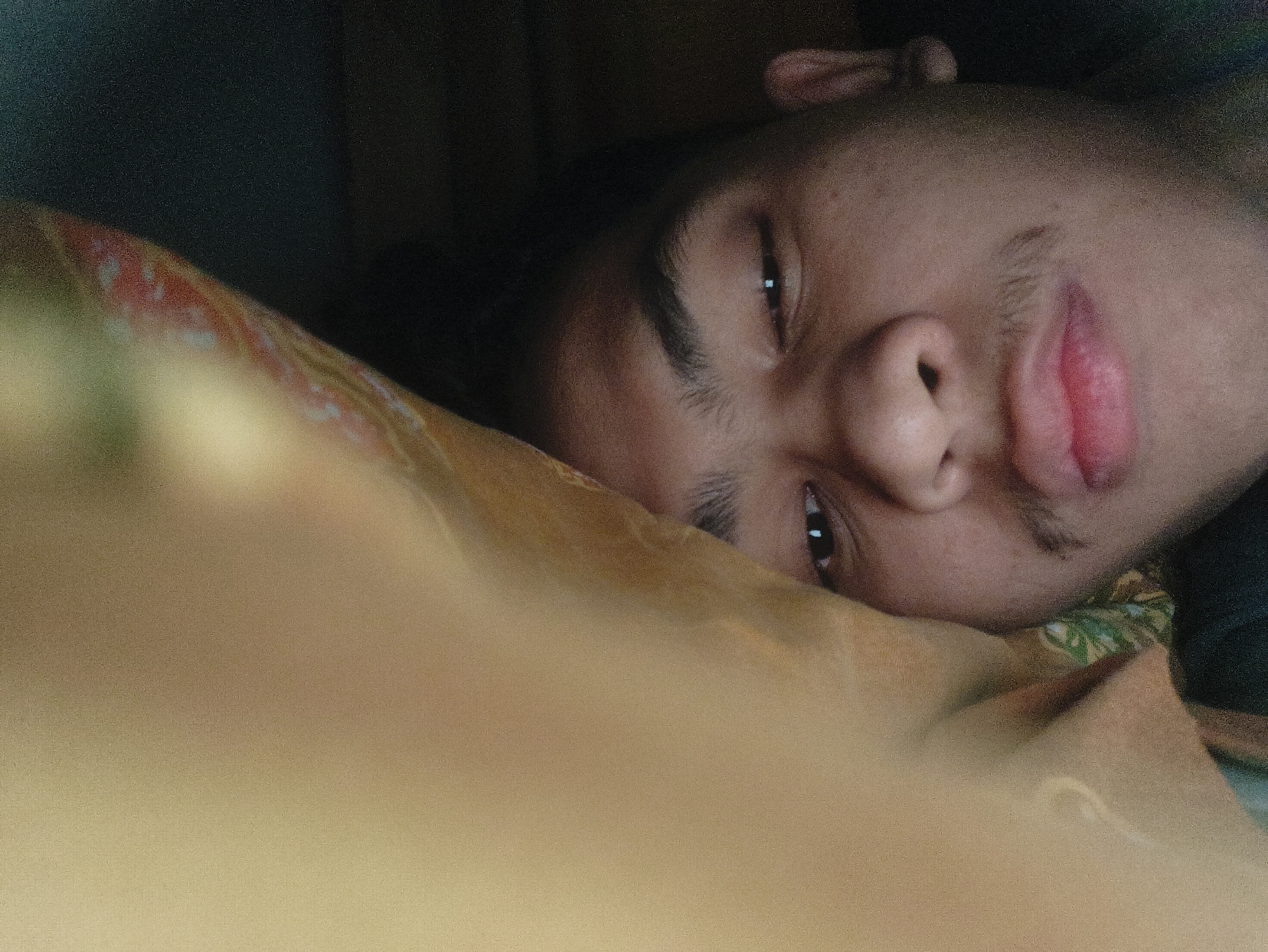 A young male individual with medium brown skin tone, dark hair, and faint facial hair (mustache and chin area) is depicted in a close-up, horizontal orientation. He is lying on his right side, with his head resting on a patterned pillow or bedding. The pillow fabric is light brown/tan with subtle patterns in muted green, red, and possibly light blue. The subject's eyes are open, looking directly towards the viewer, with light reflections visible in the pupils. His lips are slightly parted, revealing a pinkish lower lip, and a subtle smirk is present. The upper left and top portions of the background are dark and out of focus, suggesting a dimly lit indoor environment. A dark fabric, possibly clothing, is visible behind his head on the left. The foreground, closest to the camera, is an out-of-focus blur of light brown fabric. The specific city of Lhokseumawe, Indonesia, cannot be determined from the visual evidence within the image.