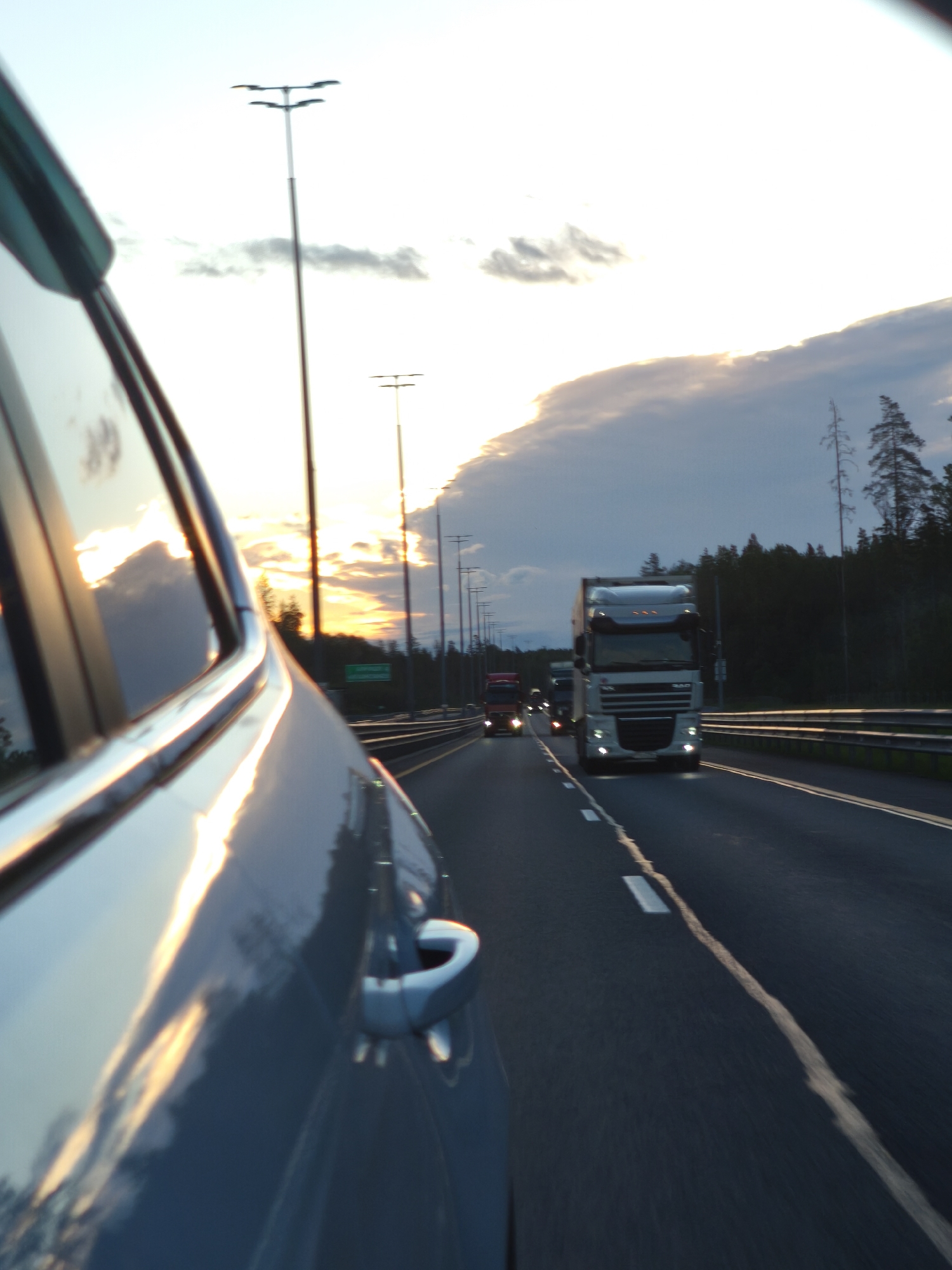 The image depicts a highway scene at dusk, possibly in or near Ushaki, Russia. The perspective is from inside a moving vehicle, with the side of a blue car taking up the left foreground. The car's reflective surface shows the sunset and clouds. On the highway, several trucks are visible, with a white semi-truck in the center lane prominently featured. Streetlights line the side of the road, and the sky is a mix of blue and orange hues from the setting sun, with scattered clouds. The environment appears to be a wooded area bordering the highway.