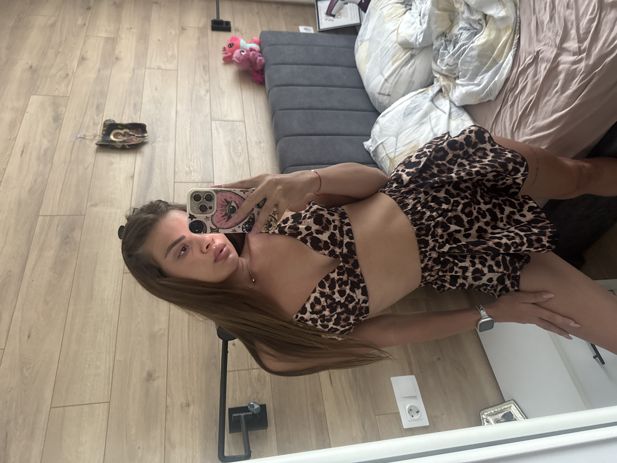 Here is a description of the image:
A woman is taking a selfie in a mirror. She is wearing a leopard print two-piece outfit. The background shows a room with wooden floors, a bed with a gray headboard, white and brown sheets, and some other objects scattered around. These include a small pink stuffed animal and a few other miscellaneous items. A floor lamp and a small picture frame are visible near the mirror.