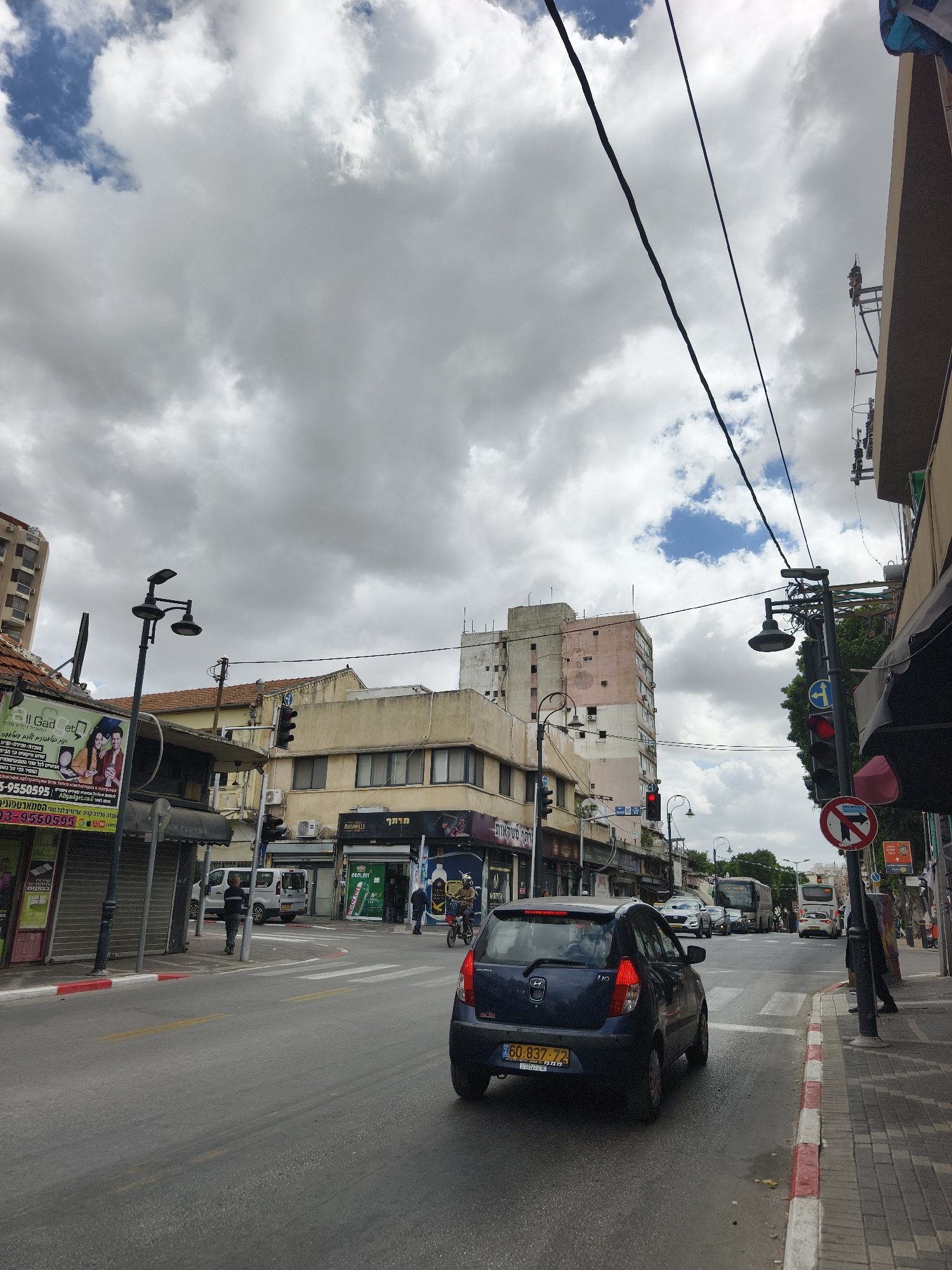 The image depicts a street scene in Petah Tikva, Israel. A blue car is in the foreground, stopped at a crosswalk and facing a red traffic light. The road is asphalt and appears to be slightly wet. On either side of the road, there are buildings with shops at street level. Storefronts include "All Gadget", and "BUSHMILLS" with Hebrew signage. Several pedestrians are present on the sidewalks and crossing the road. There are lamp posts and power lines overhead, and the sky is overcast with heavy clouds. The license plate of the car reads "60-837-72".