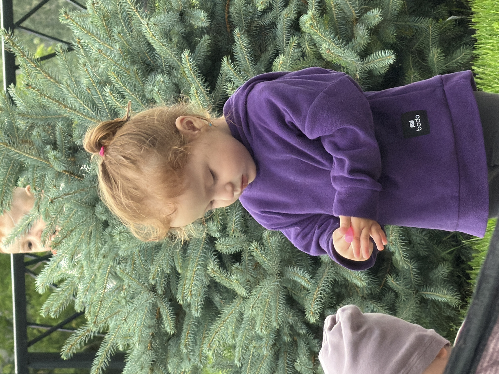 A young child with reddish-blond hair pulled into a ponytail stands in front of a large evergreen tree. The child is wearing a purple hoodie and looking down at their hands. There is another child in the foreground wearing a grey beanie. The scene appears to be outdoors, possibly in a park or backyard in Tashtagol, Russia.