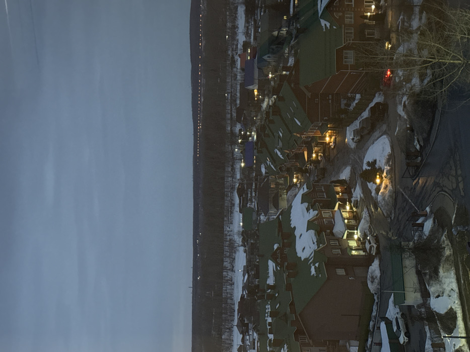 This image presents an elevated, static view of a residential neighborhood in Chaikovsky, Russia, observed during dusk or early night in a winter setting. The sky is a uniformly dark blue-grey, suggesting an overcast condition. On the distant horizon, a dark, tree-lined landmass is visible, punctuated by a continuous horizontal line of yellow artificial lights, indicative of a road or industrial infrastructure. Below this, a frozen or snow-covered body of water separates the distant land from the immediate residential area.
The foreground and midground feature a clustered development of two-to-three-story residential buildings, predominantly constructed with reddish-brown brick or siding and characterized by dark green pitched roofs, many of which retain patches of snow. Numerous windows and entryways are illuminated with warm yellow light. A network of wet, dark asphalt roads crisscrosses the development, bordered by snow accumulations. A circular street island, possibly a small roundabout, is centrally located within the residential cluster and features an illuminated evergreen shrub.
Several vehicles are present: a white SUV is parked on a snow-covered verge in the lower-left. Another white SUV is parked in front of a building further up on the left side of the street. In the center-right, a light-colored sedan and a dark vehicle are parked on the street. Further right, a red vehicle with illuminated taillights is visible on the roadway. No individuals are discernible within the scene. The environment indicates cold weather conditions with substantial snow cover on non-road surfaces.