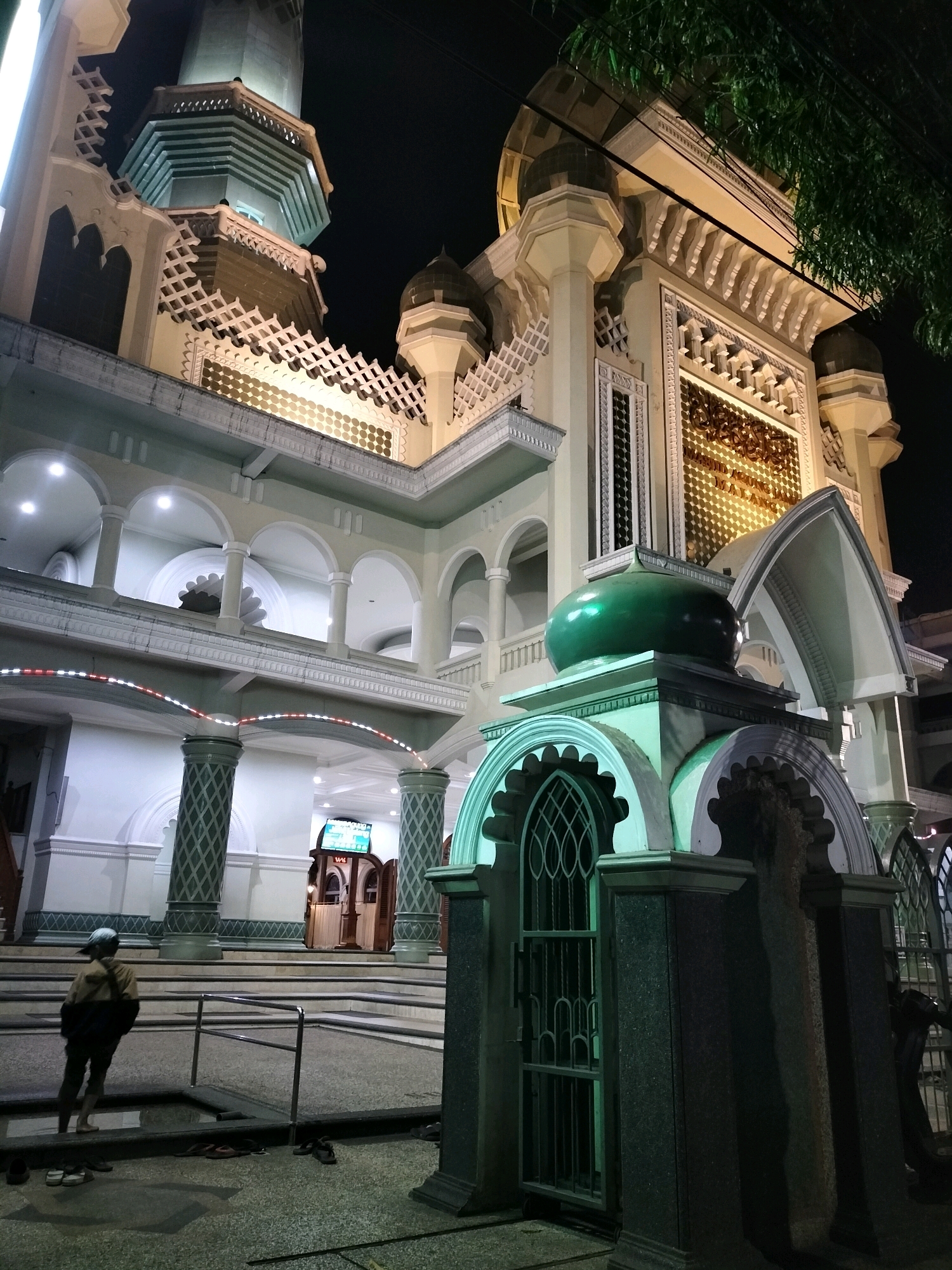 The image captures the facade of a mosque at night. The mosque has multiple levels with arched windows and decorative elements, illuminated by artificial lighting. A tall, octagonal minaret rises from the structure, also lit from below. In the foreground, a small, green-domed structure with intricate metalwork stands. To the left, a person wearing a cap and a jacket with a backpack is standing near a metal railing. The ground is paved with speckled stone, and several pairs of footwear are scattered around. The overall scene suggests a nighttime view of a religious building in an urban setting. The location of the mosque is Masjid Agung Jami' Malang, located in the city of Malang, Indonesia.