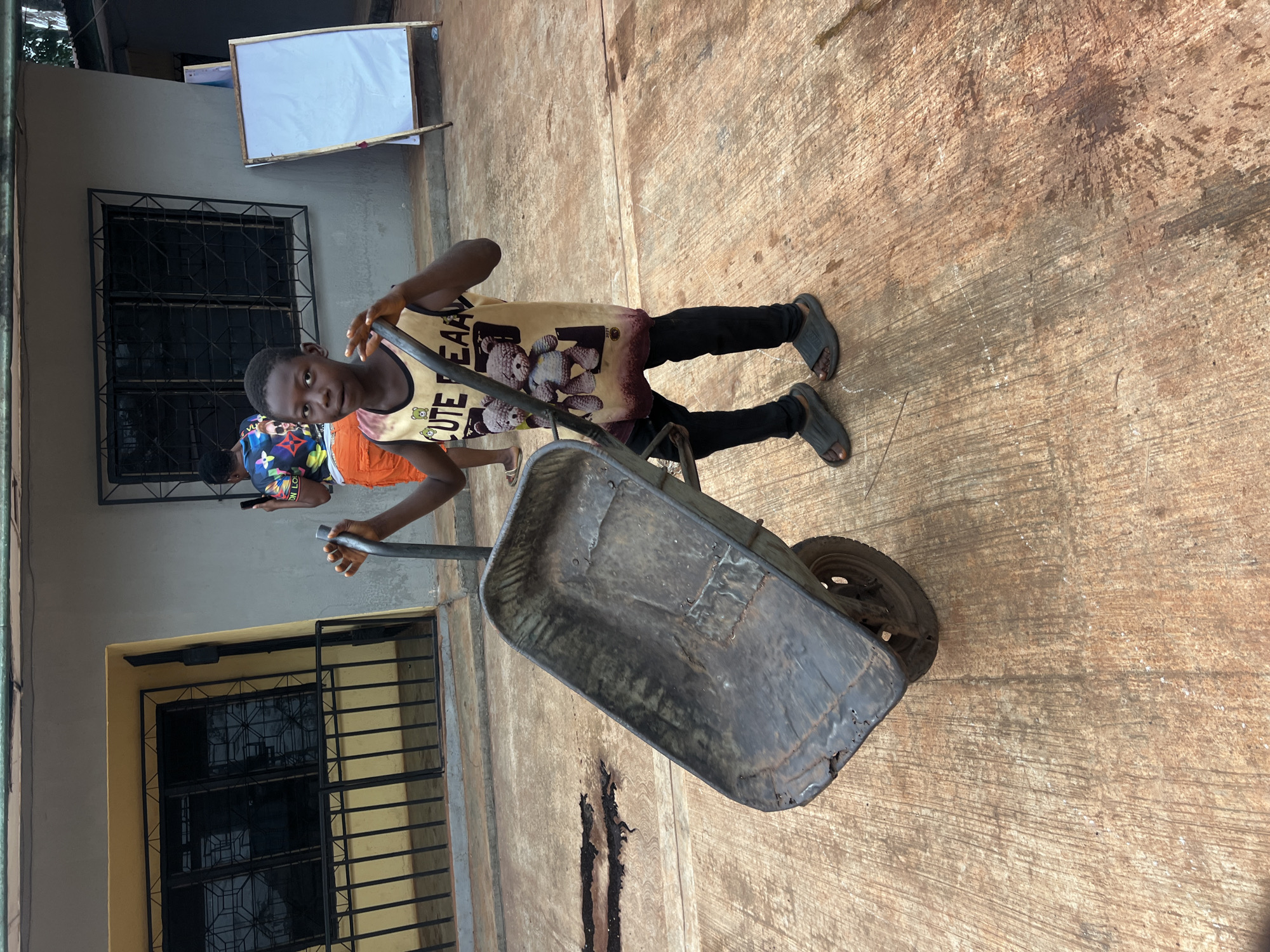 An outdoor scene, likely in Onitsha, Nigeria, features a young person holding a metal wheelbarrow. The main subject, a young male, is positioned in the foreground, wearing a light-colored tank top adorned with the text "CUTE BEAR" and a teddy bear graphic, black trousers, and dark slip-on sandals. He is holding the handles of an aged, rusty wheelbarrow, which shows considerable wear and tear, and is looking directly towards the camera with a neutral expression.
In the background, another person is partially visible, dressed in a colorful patterned shirt and orange shorts, holding a mobile phone. They are standing near a building with multiple black barred windows, a common architectural feature in the region. A white board with a wooden frame is propped against the building wall.
The ground is a rough, discolored concrete or paved surface, showing cracks and stains. The lighting is bright and even, indicating daytime conditions, possibly midday or early afternoon, under clear skies. The scene suggests a functional area, potentially a yard or work site, reflecting everyday activities.