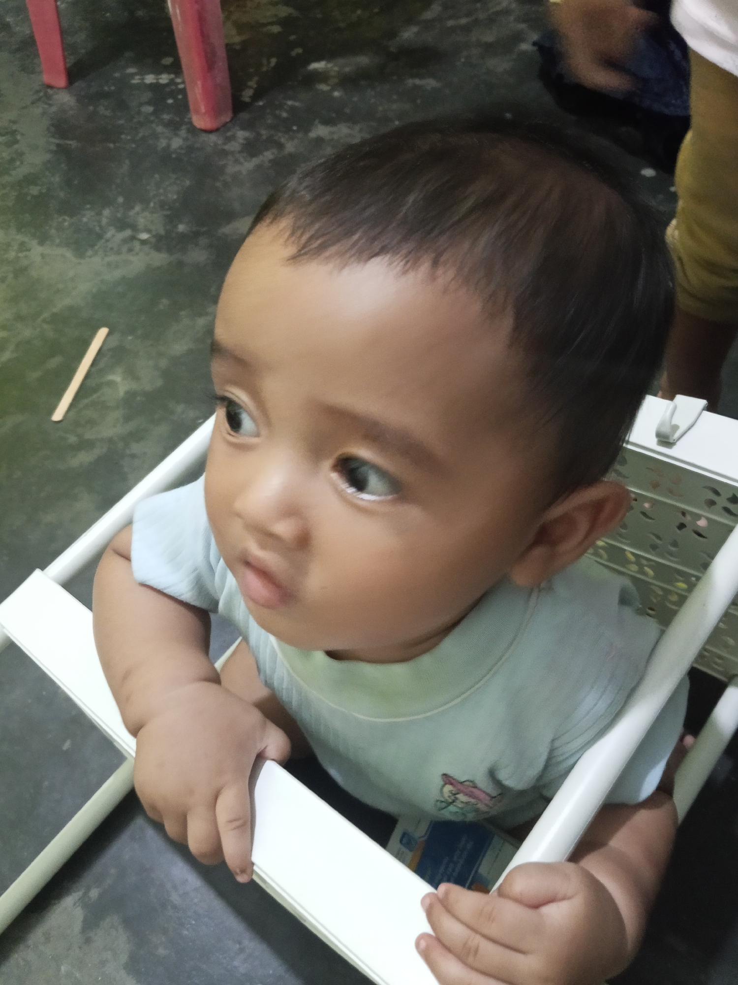 An overhead, medium shot shows a toddler sitting in a white chair. The child is facing right, with their head turned to look to the left. The toddler has dark hair, brown eyes, and light brown skin. They are wearing a light blue t-shirt. The child's left arm is resting on the chair, and their right hand is also on the chair. The background is a dark, concrete floor, with parts of red chairs visible in the upper left corner. A small, light brown stick is on the floor to the left of the child. The location is identified as Lestari Indah, Indonesia.