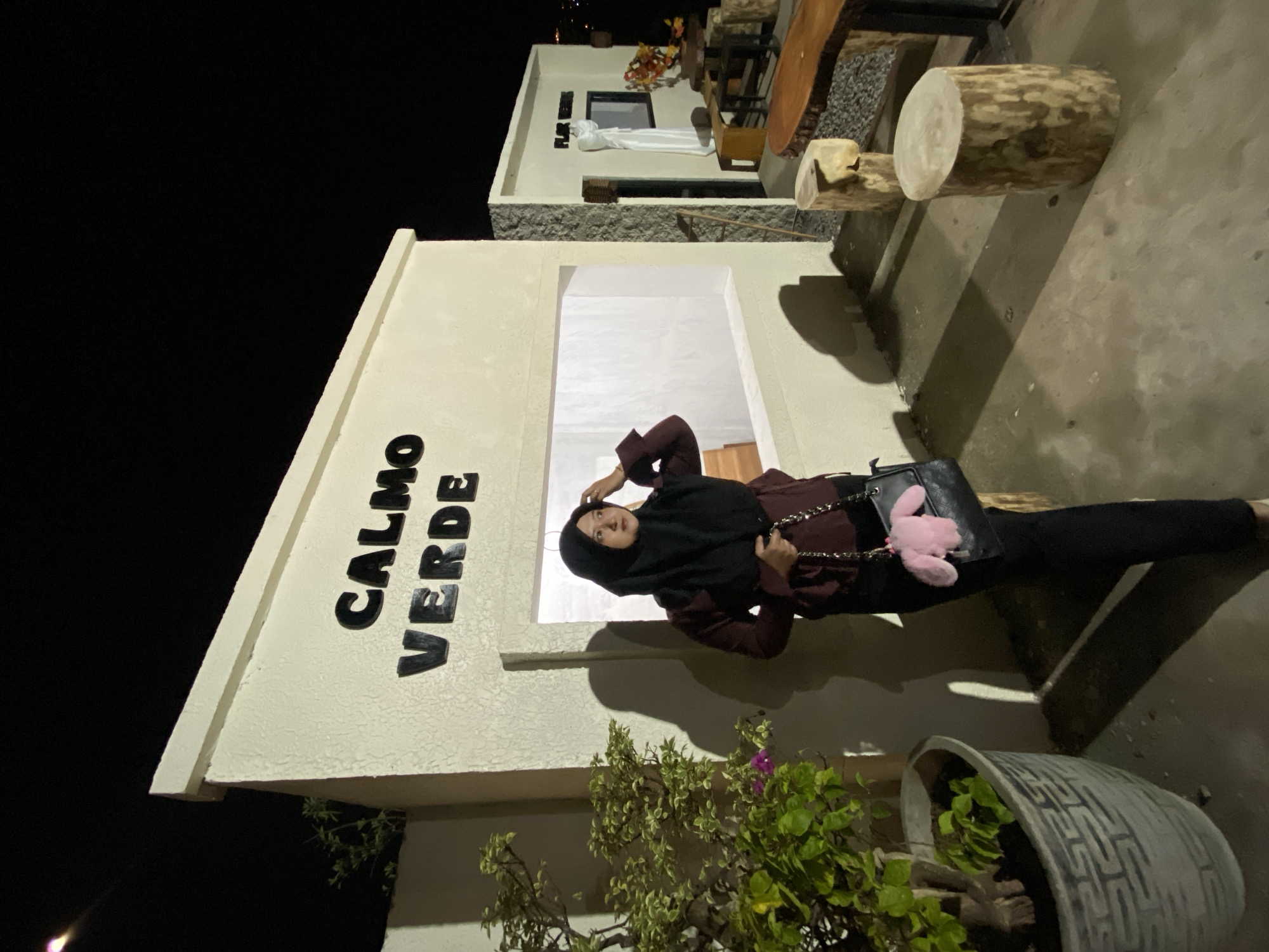 A medium shot captures a woman in a black hijab and a burgundy long-sleeved top, standing in front of a cream-colored building with the words "CALMO VERDE" in black, raised lettering on its facade. She is holding a black purse with a pink fuzzy charm. To her left is a large potted plant with green and variegated leaves. The ground is concrete. In the background, there is a smaller building with "FLOR VERDE" signage and an outdoor seating area with log-style stools and wooden tables. The scene appears to be at night, as indicated by the dark sky and shadows cast by artificial lighting. The image was taken in the city of Kesiman, Indonesia.