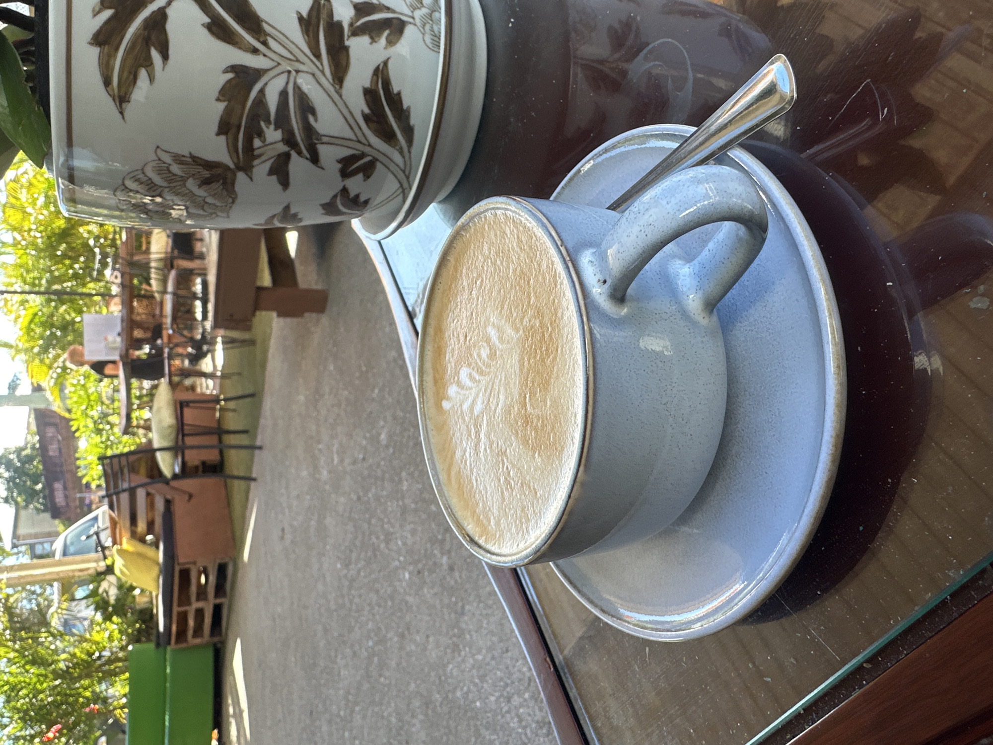 A close-up shot captures a light blue ceramic mug of latte art resting on a matching saucer. A silver spoon lies across the saucer. The mug and saucer are positioned on a dark, reflective glass-topped table. To the right, a portion of a ceramic pot decorated with brown floral patterns is visible. In the background, the scene opens to an outdoor seating area of a cafe, with various tables and chairs. Sunlight filters through lush green foliage, suggesting a bright day. A glimpse of a person can be seen in the distance, seated at a table. The environment suggests a relaxed cafe setting, likely in Bellingen, Australia, given the implied presence of outdoor seating and lush greenery characteristic of the region.