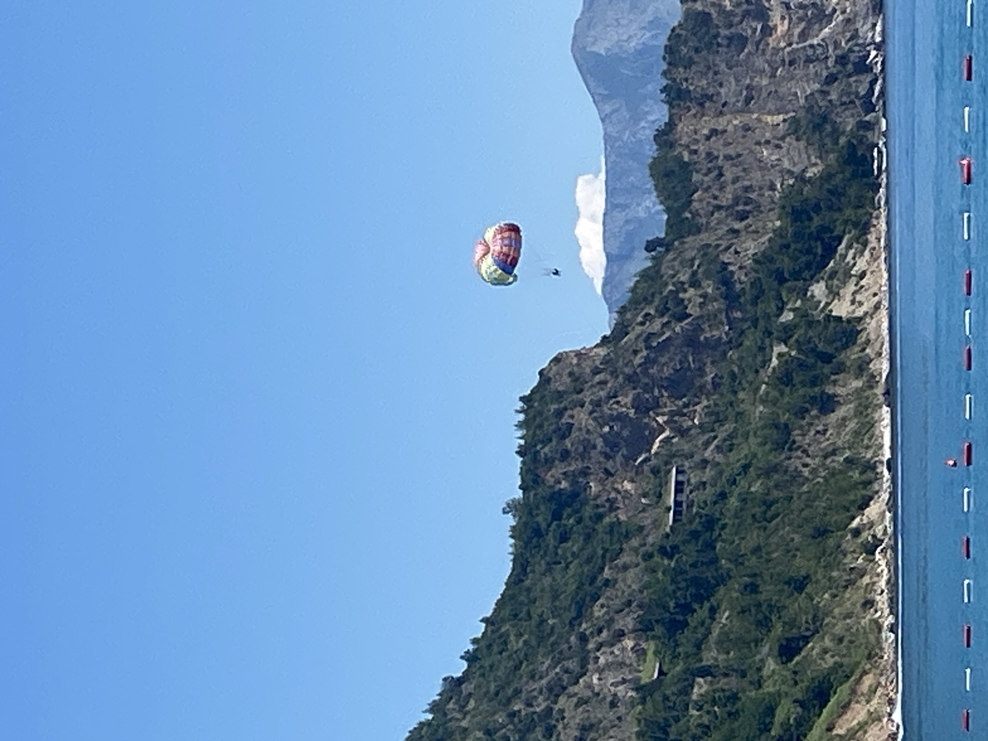 This media file depicts a paraglider soaring through a clear blue sky, with a rugged, tree-covered mountain and a calm body of water in the background. The paraglider, a colorful canopy of red, yellow, green, and blue, is the central focus in the sky. A small, dark silhouette of a person can be seen beneath the paraglider. The mountain slopes are densely vegetated, with patches of exposed rock. The water, to the right of the frame, is a deep blue and appears to have a series of white and red buoys marking an area. The image suggests a bright, sunny day, likely during daylight hours, given the clear sky and strong lighting. The scene evokes a sense of adventure and freedom associated with paragliding, set against the dramatic natural landscape of Prijevor, Montenegro.