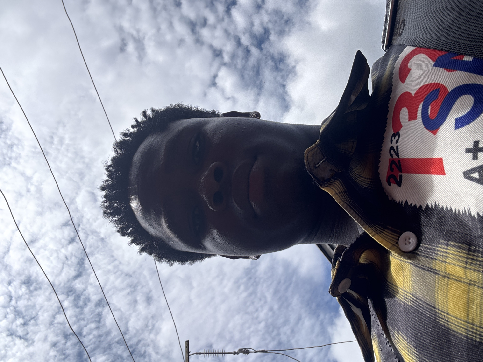 A low-angle, full-frame, eye-level, outdoor, close-up, portrait photograph captures a young Black male from the chest up against a cloudy sky. He is looking directly at the camera. He has short, dark, curly hair and is wearing a yellow, black, and white plaid shirt. The shirt has a prominent white patch on the right side of the chest featuring large red and blue lettering that reads "2223 TS A+". The photograph appears to have been taken in the city of Ihiagwa, Nigeria, given the overcast, humid sky and the presence of overhead electrical wires. The lighting is bright, with the sun casting shadows on the subject's face, particularly around his nose and under his chin. The background is dominated by a sky filled with scattered, white, cumulus clouds. A thin antenna-like object is visible on the left side of the frame, attached to what appears to be a building.