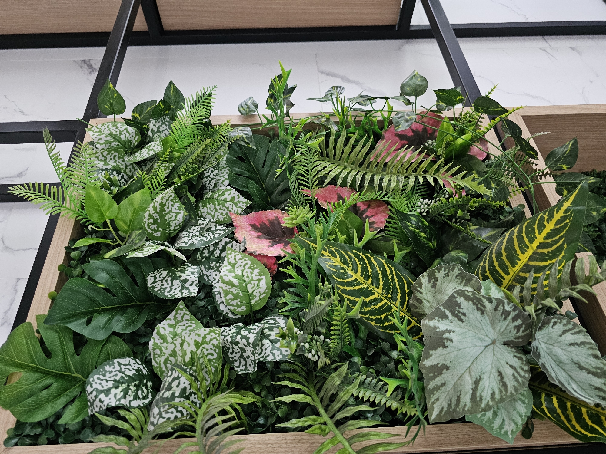 This is a close-up, eye-level, medium shot of a section of a decorative vertical garden wall constructed from artificial plants. The garden is housed within a light-colored wooden frame. The wall is densely packed with a variety of artificial foliage in shades of green, white, yellow, and pink. Prominent leaf shapes include large, lobed green leaves (likely Monstera), smaller leaves with white variegation and speckles (possibly artificial Dieffenbachia or similar), delicate ferns, and broader leaves with striking yellow and green striped patterns. Some pink-tinged leaves with dark green veins are also visible. The background consists of a white marbled wall and a black metal shelf structure. The lighting is bright and even, suggesting an indoor setting. There are no people or animals present in the image. The overall impression is one of lush, well-maintained artificial greenery.