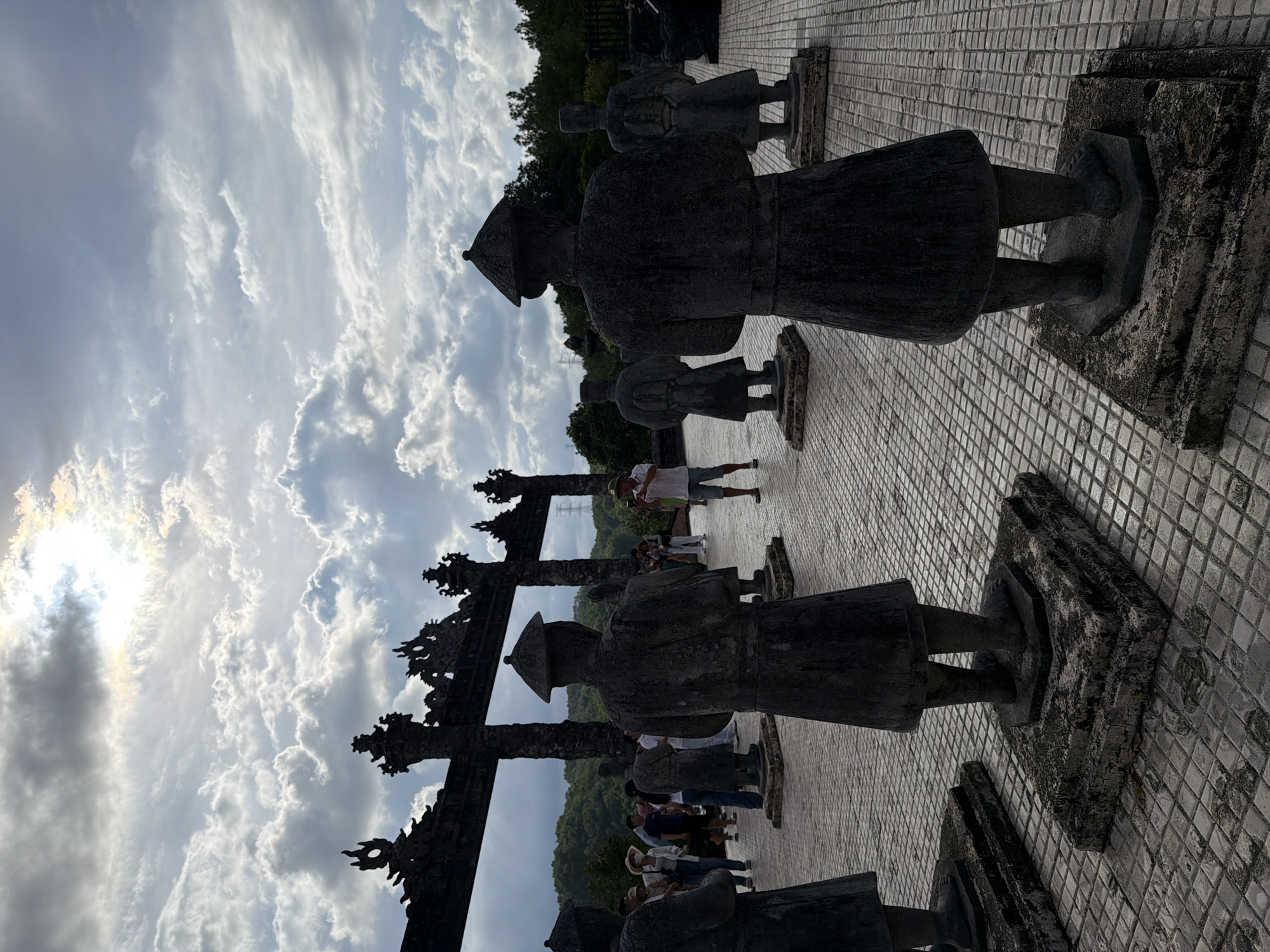 In Thủy Xuân Ward, Vietnam, a scene unfolds beneath a dramatic, cloud-filled sky with the sun peeking through. The foreground is dominated by the backs of four dark, stone statues, each standing on a tiered, moss-covered pedestal. These statues, likely of historical figures, wear traditional conical hats and long robes, their stances solemn and unmoving.
Behind the statues, a large, ornate stone gateway, also dark and imposing, stands as a backdrop. The gateway features intricate carvings and is flanked by stone columns. In the middle ground, several tourists are visible, some walking, others observing. One man in a white shirt and blue shorts stands on the tiled ground, holding up his phone, seemingly taking a photograph. Other tourists, dressed in casual attire, are scattered throughout the area, some appearing to chat or pose for pictures.
The ground is paved with small, square tiles, arranged in a grid pattern. Beyond the gateway, lush green trees and rolling hills form the distant landscape. The overall atmosphere suggests a historical site, likely a temple or imperial tomb, attracting visitors to explore its ancient architecture and cultural significance. The interplay of the strong sunlight and the heavy clouds creates a somewhat moody and awe-inspiring ambiance.