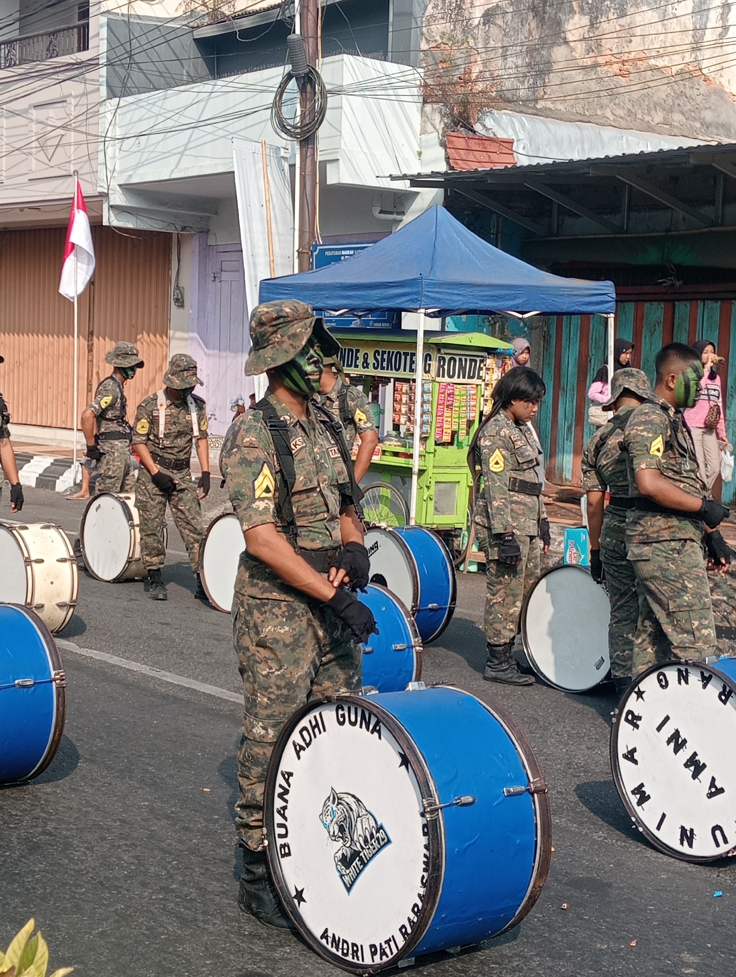 This image captures a street scene in Bintoro, Indonesia, likely during a parade or public event. In the foreground, several individuals in camouflage military uniforms stand with large drums. The drums are a mix of white and blue, with some featuring white tiger logos and text such as "BUANA ADHI GUNA" and "UNI ARMINO RAPA". The individuals are wearing hats, gloves, and some have face paint in green and black. One person in the center is looking down at a drum, while others appear to be in formation. In the background, there are buildings, a blue tent, and a food cart. An Indonesian flag is visible on a pole to the left of the frame. The overall atmosphere suggests a festive or organized procession.