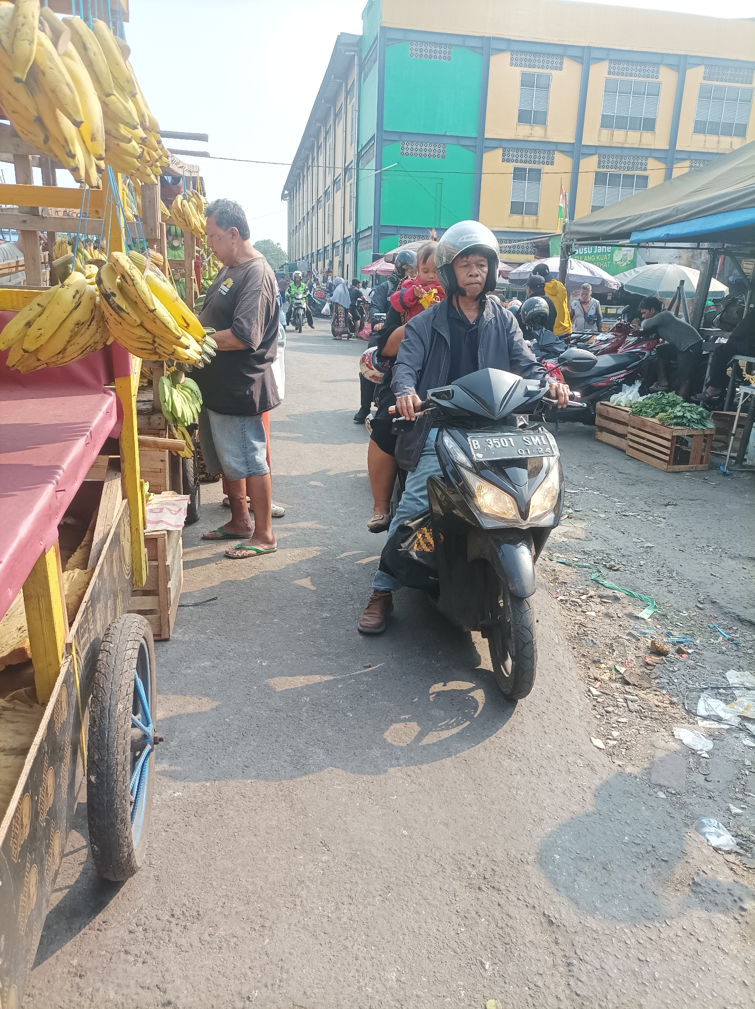 This is a street scene in South Jakarta, Indonesia, with a focus on a market area. In the foreground, a man wearing a helmet rides a black scooter with a passenger behind him, a child. The scooter has a license plate that reads "B 3501 SMI" and "01-24". To the left, a fruit stand displays numerous bunches of bananas hanging from a wooden structure. A man in a dark t-shirt and denim shorts stands near the fruit stand, looking at the bananas. Further back, other people are visible, along with more market stalls and buildings. The environment appears to be a busy outdoor market with concrete pavement and some debris on the ground. The buildings in the background are colorful, with yellow and green sections, suggesting commercial establishments. Sunlight indicates it is daytime.