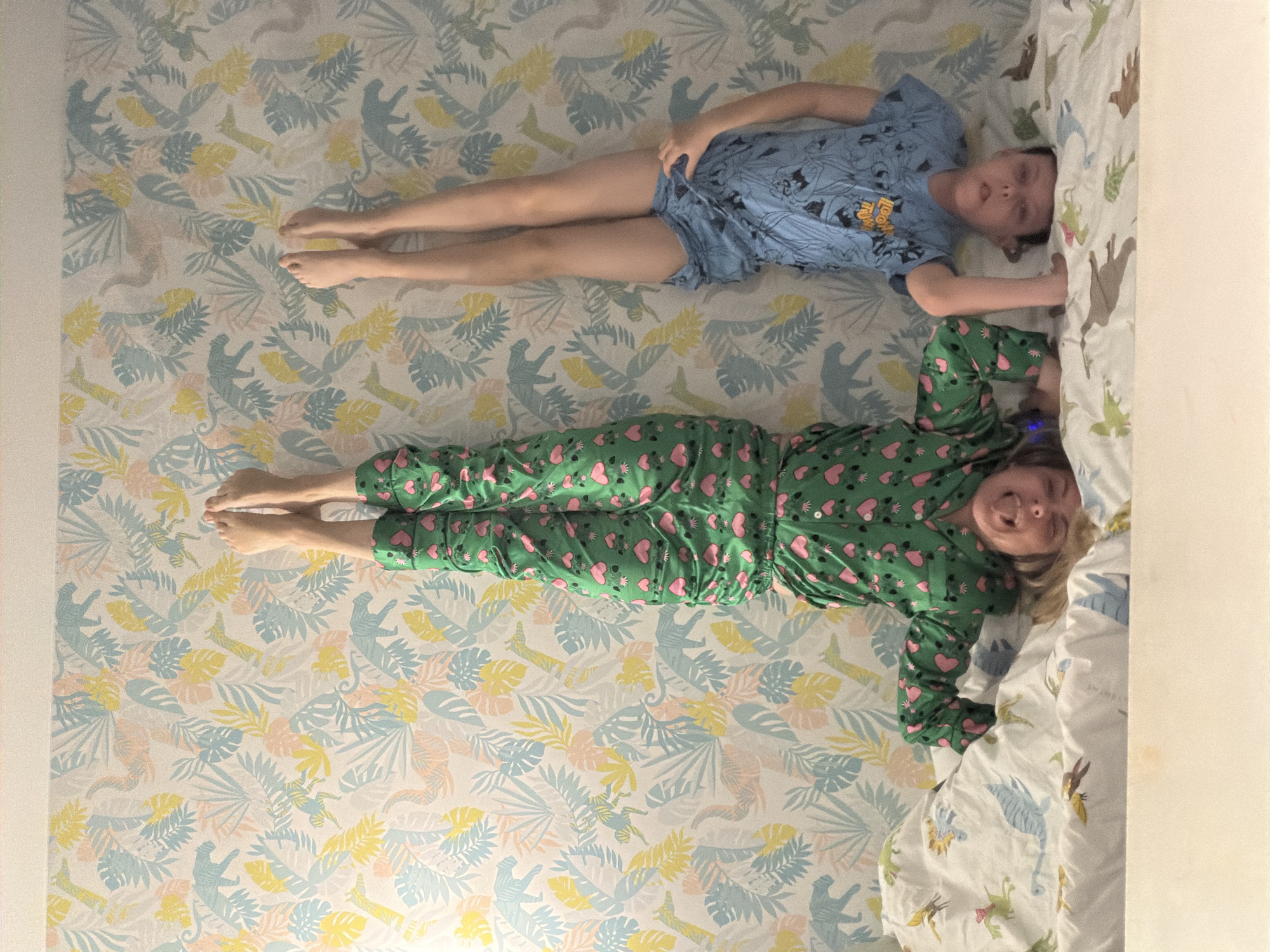 An overhead shot taken in Votkinsk, Russia, shows two children lying on a bed covered with a light-colored sheet featuring a pattern of pastel yellow, light blue, and peach tropical leaves and dinosaur silhouettes.
On the left, a child wearing a blue t-shirt and shorts lies flat on their back, with legs extended and arms resting at their sides. The blue t-shirt has a graphic print, and the text "LOONEY TUNES" is partially visible across the chest. This child looks towards the camera with a neutral expression.
On the right, closer to the camera, another child is lying on their back, wearing green pajamas covered in pink heart patterns. This child has blonde hair and is smiling broadly at the camera. Their legs are extended, and their arms are bent, resting on the bed.
A white duvet cover with illustrations of various dinosaurs in brown, green, and blue is visible on the right side of the bed. A plain white wall runs along the far left edge of the frame. Both children appear relaxed and content, posing for the photograph in an indoor setting, likely a bedroom. The lighting is soft and even.