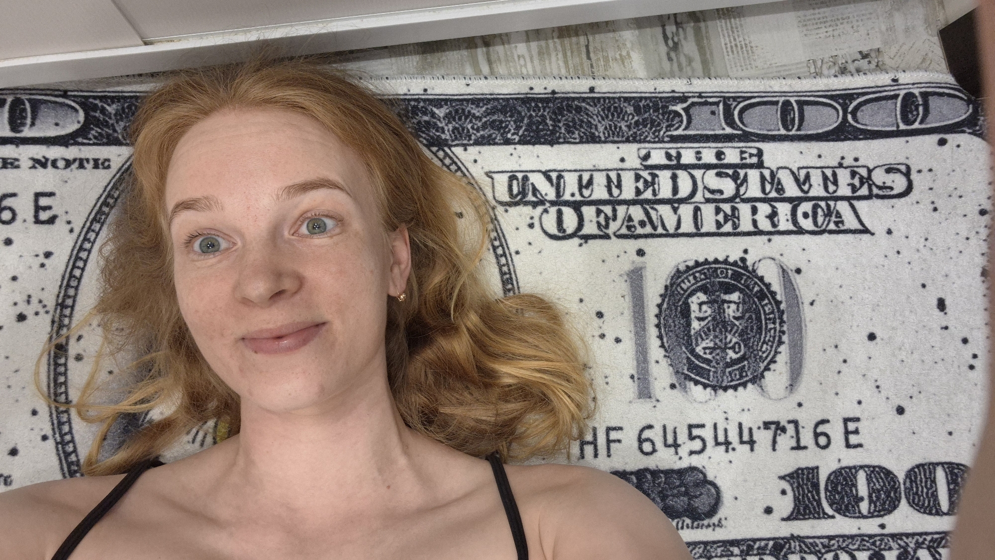 A forensic description of the scene reveals a single female individual positioned supine on a patterned surface. The individual, identified as a woman, possesses long, wavy, reddish-blonde hair spread around her head. Her eyes are light blue-green and directed toward the camera with a wide gaze. Her complexion is fair with visible freckles, and her lips are slightly parted in what appears to be a subtle smile. A small, simple gold hoop earring is visible in her right ear. She is wearing a black spaghetti strap garment, visible on her left shoulder and upper chest.
The patterned surface beneath her is a rug or blanket designed to mimic an oversized United States $100 bill. Visible details on the pattern include "UNITED STATES OF AMERICA," a stylized "100" denomination, portions of "ONE HUNDRED DOLLARS," and a serial number reading "HF 64544716E." The pattern incorporates white, black, and gray tones and features dark speckles simulating wear or design. A narrow, plain white horizontal strip, indicative of an interior architectural feature such as a wall or ceiling element, is visible above the woman's head. The scene is illuminated by ambient indoor light. The location is the city of Chaikovsky, Russia.
