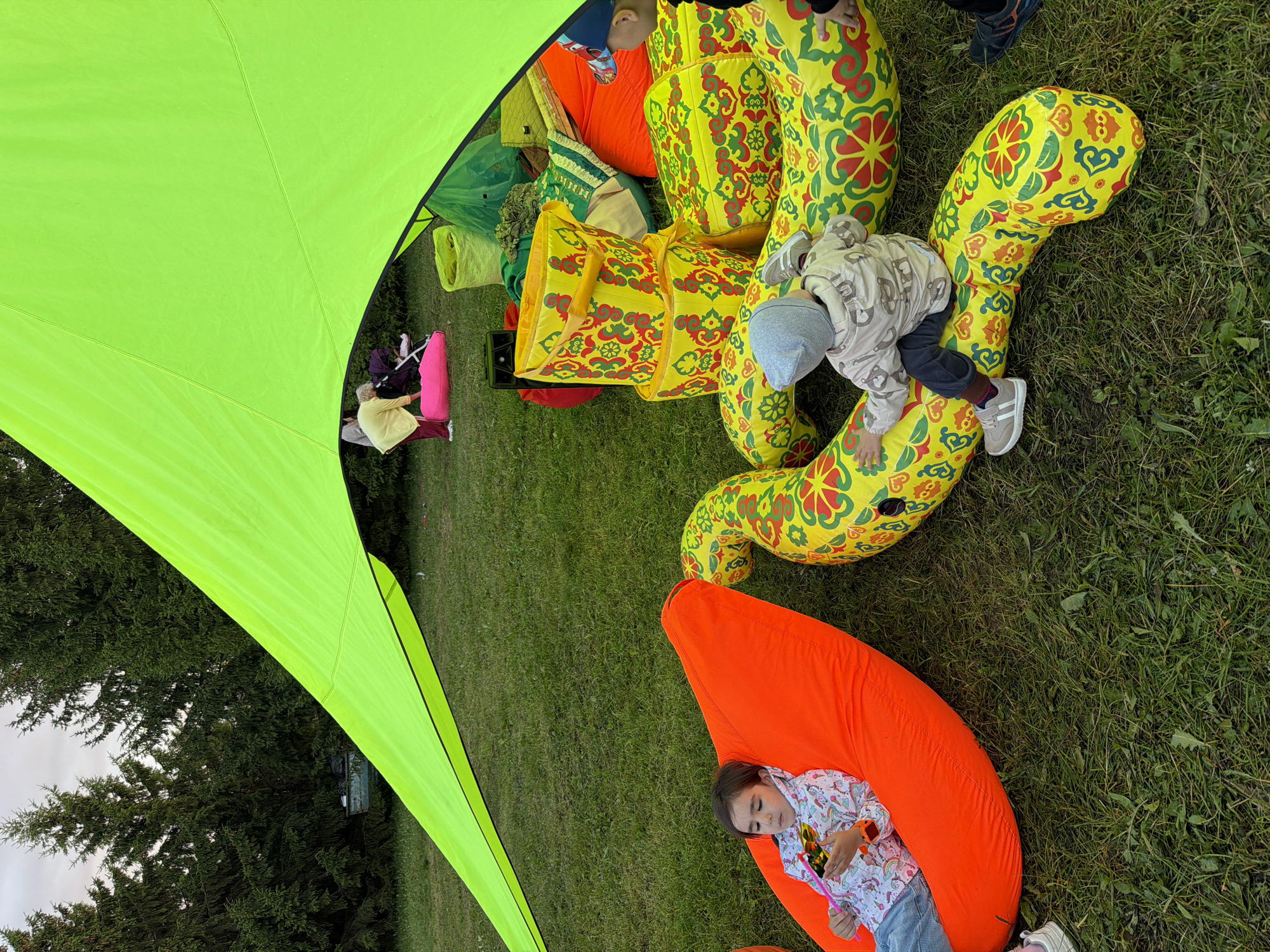 In an outdoor setting, under a bright neon green tarp, a group of people are gathered on a grassy area. A young girl with dark hair is seated on a large, bright orange beanbag chair, holding a toy. Next to her, a child wearing a grey hat and a patterned hooded sweatshirt is sitting on a long, inflatable yellow cushion adorned with a red and green floral pattern. In the background, several adults are visible, along with a stroller and various colorful inflatable objects and bags. The environment appears to be a park or similar recreational space, with trees in the distance under a cloudy sky.