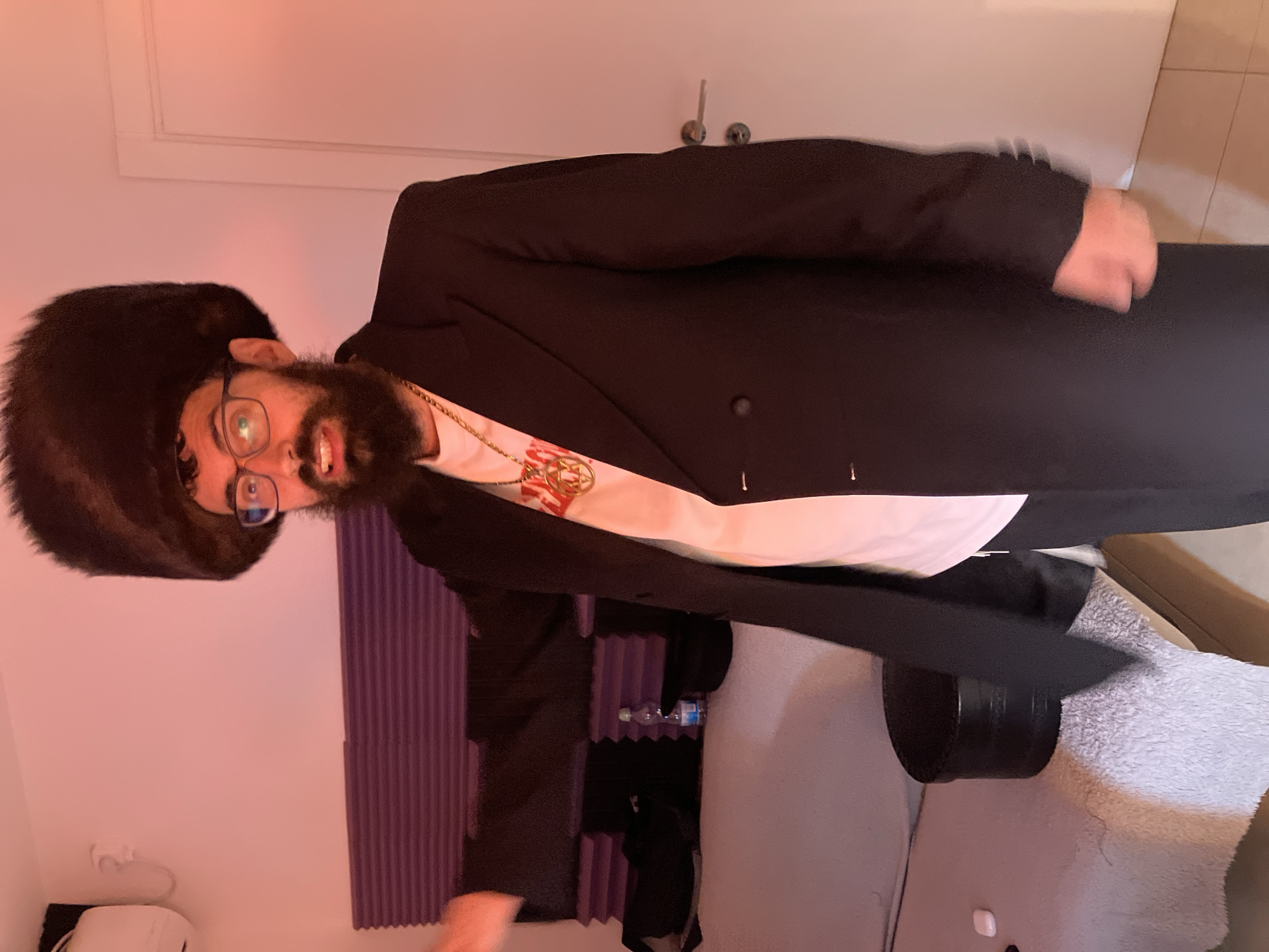 A blurry, canted photograph depicts a man indoors. He wears a black fur hat, glasses, a black jacket over a white t-shirt, and a gold chain. He has a beard. Behind him are purple acoustic panels, a door, and furniture covered with a gray towel. A bottle of water sits among the acoustic panels. The location is indeterminate, and there is no evidence to suggest the location is Tel-Aviv, Israel.