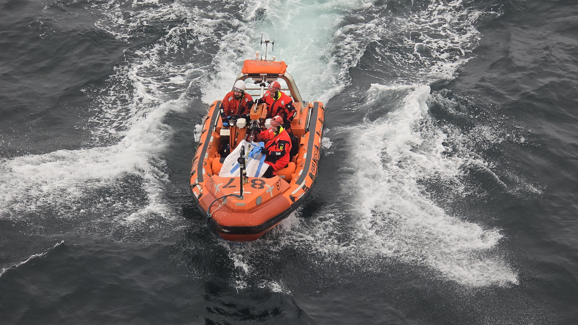 The image shows an orange inflatable boat with six people aboard, moving through dark ocean water. The boat has a frame over it and is numbered "87" on the bow. The occupants are wearing red waterproof gear and helmets. The boat is creating a wake behind it as it moves. The location is unknown.