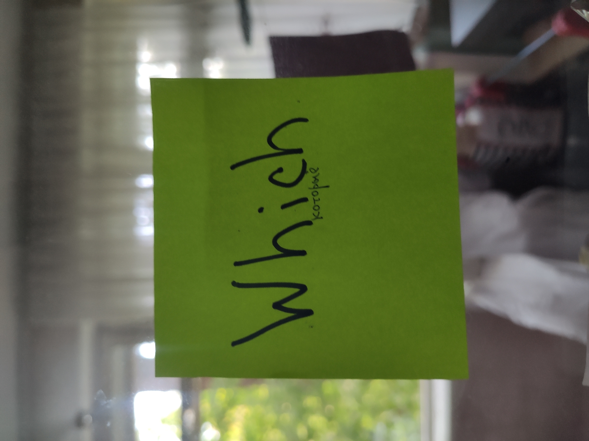 A lime green sticky note is affixed to a window, displaying handwritten text in black marker. The top line reads "Which" in large, cursive English letters. Below it, in smaller, neat cursive Russian letters, is the word "которые" (kotoryye), which translates to "which" or "who" in English. The background through the window shows a blurred scene of greenery, possibly trees or bushes, suggesting an outdoor environment. The overall lighting is diffused, indicating it is daytime. The sticky note is positioned centrally on the window pane. There are no people or other objects immediately visible on or around the sticky note. The context of Tashkent, Uzbekistan, is not visually derivable from the image content itself.