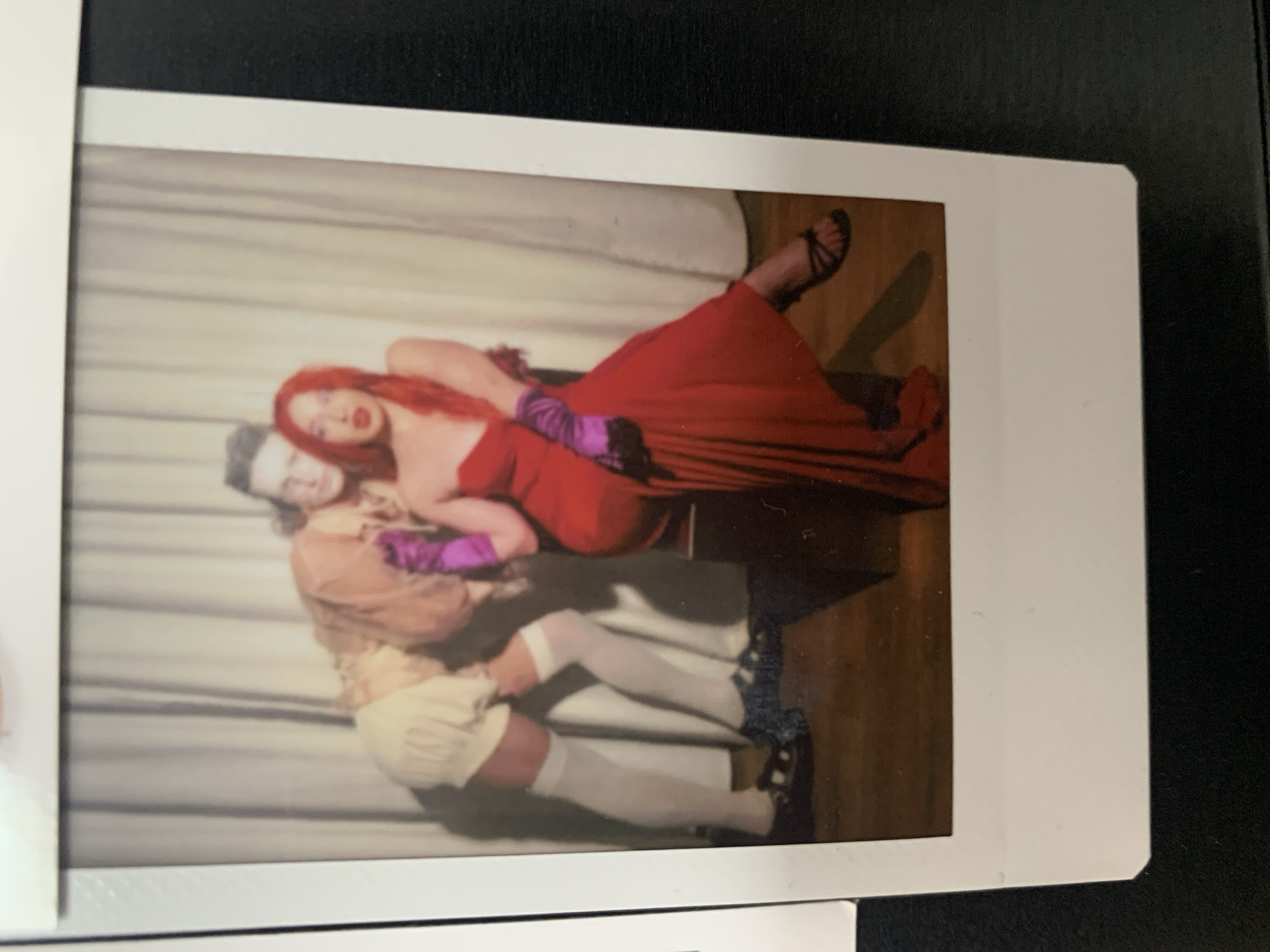 The image is a Polaroid photograph depicting two people in costume. A person with bright red hair and wearing a strapless red dress and purple gloves reclines on a surface or chair, with black strappy sandals visible on their feet. Another person, dressed as Roger Rabbit in a brown shirt, tan shorts, white stockings, and black shoes, is also visible. They are posing together. The background appears to be a draped sheet or curtain. The location cannot be identified as Tel-Aviv or anywhere else based on visual information.