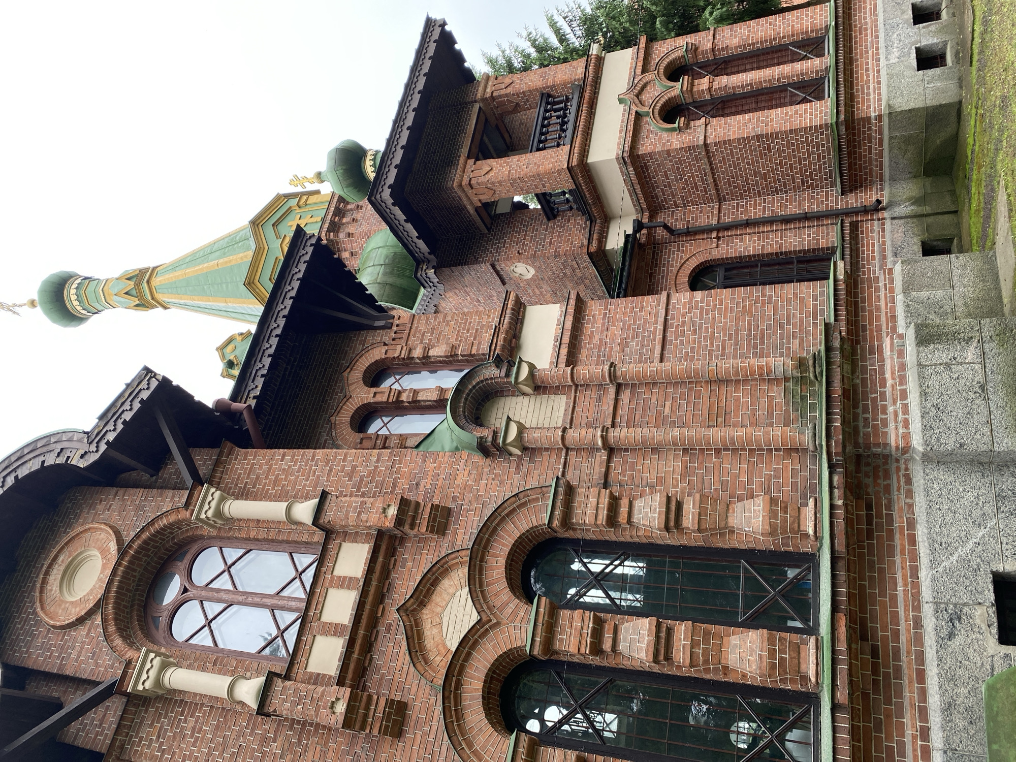 This is an exterior, medium-angle, eye-level, daylight, full-color photograph of a red brick building, possibly a church or a historical building, located in Priozersk, Russia. The building features green domed roofs and a prominent green and gold spire. The architecture displays arched windows with dark frames and intricate brickwork. The facade is decorated with pilasters and ornate details around the windows and rooflines. A portion of a tree is visible on the right side of the frame, and the ground is covered with grass and some moss. The sky is overcast. There are no people or animals visible in the image.