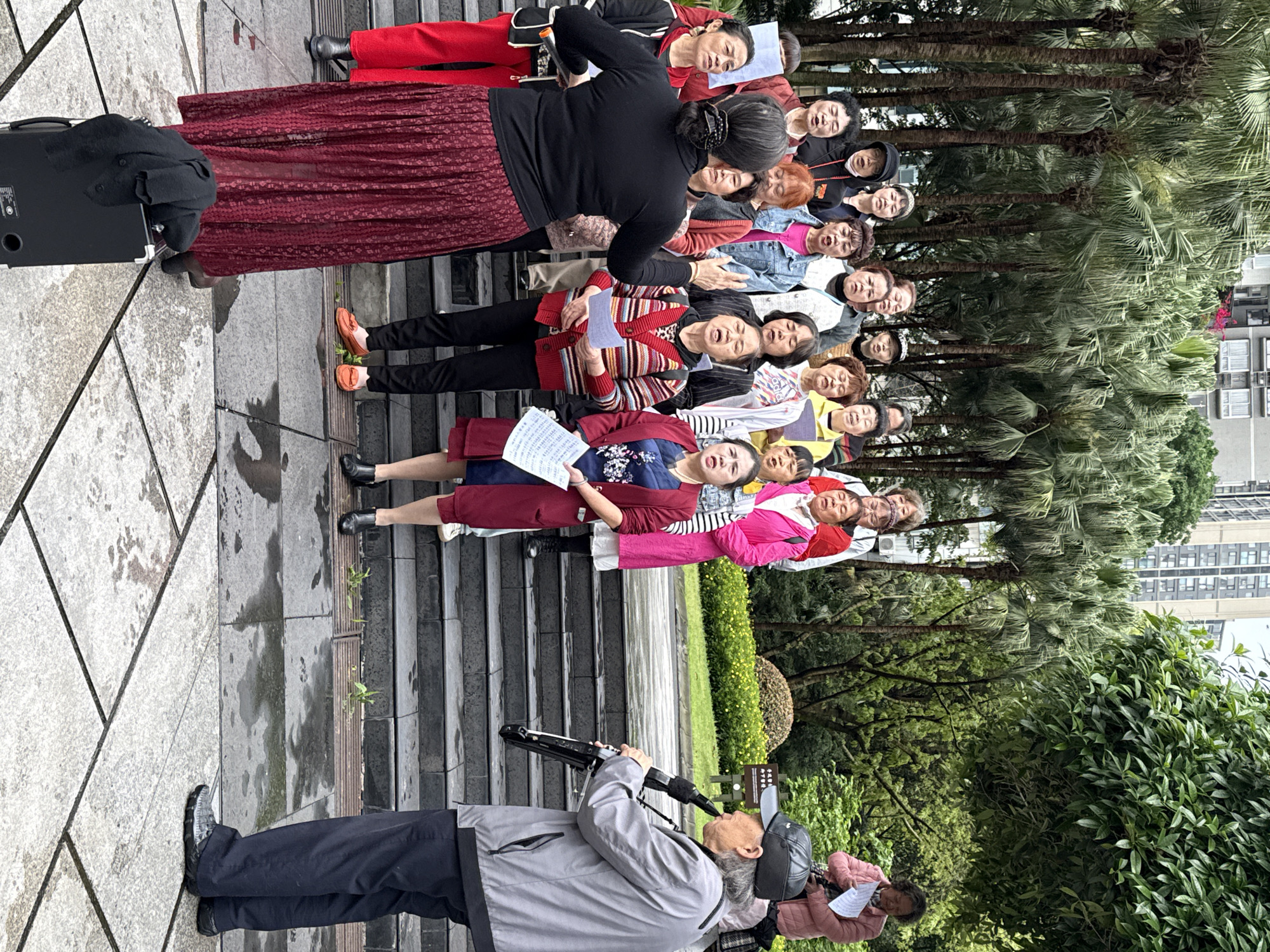 An outdoor choir, primarily composed of elderly and middle-aged individuals, is performing in what appears to be a public park or square in Jiulongpo District, China. The group, mostly women, is arranged in tiered rows on stone steps, holding song sheets and singing with open mouths and engaged expressions.
At the front of the choir, a woman in a black long-sleeved top and a long, patterned red skirt appears to be leading the performance. A black speaker or sound system is placed on the paved ground next to her. To the left, slightly separate from the main group, a man wearing a grey jacket and a black baseball cap is playing an electronic wind instrument. Another woman is seated on a bench to the far right, also holding papers.
The setting features a concrete paved area that exhibits damp patches, suggesting recent rain or morning dew. Behind the choir, several tall palm trees and other lush green trees create a natural, verdant backdrop. Distant urban buildings are visible through the foliage, indicating an urban park environment. The lighting suggests daytime under overcast or diffused conditions. Participants are dressed in casual, warm clothing like jackets and sweaters.
Visible text includes characters on the song sheets held by the performers, text on the black speaker, and partially obscured Chinese characters on a small sign positioned among the bushes in the background. The overall scene conveys a sense of community engagement and musical activity in a public green space.