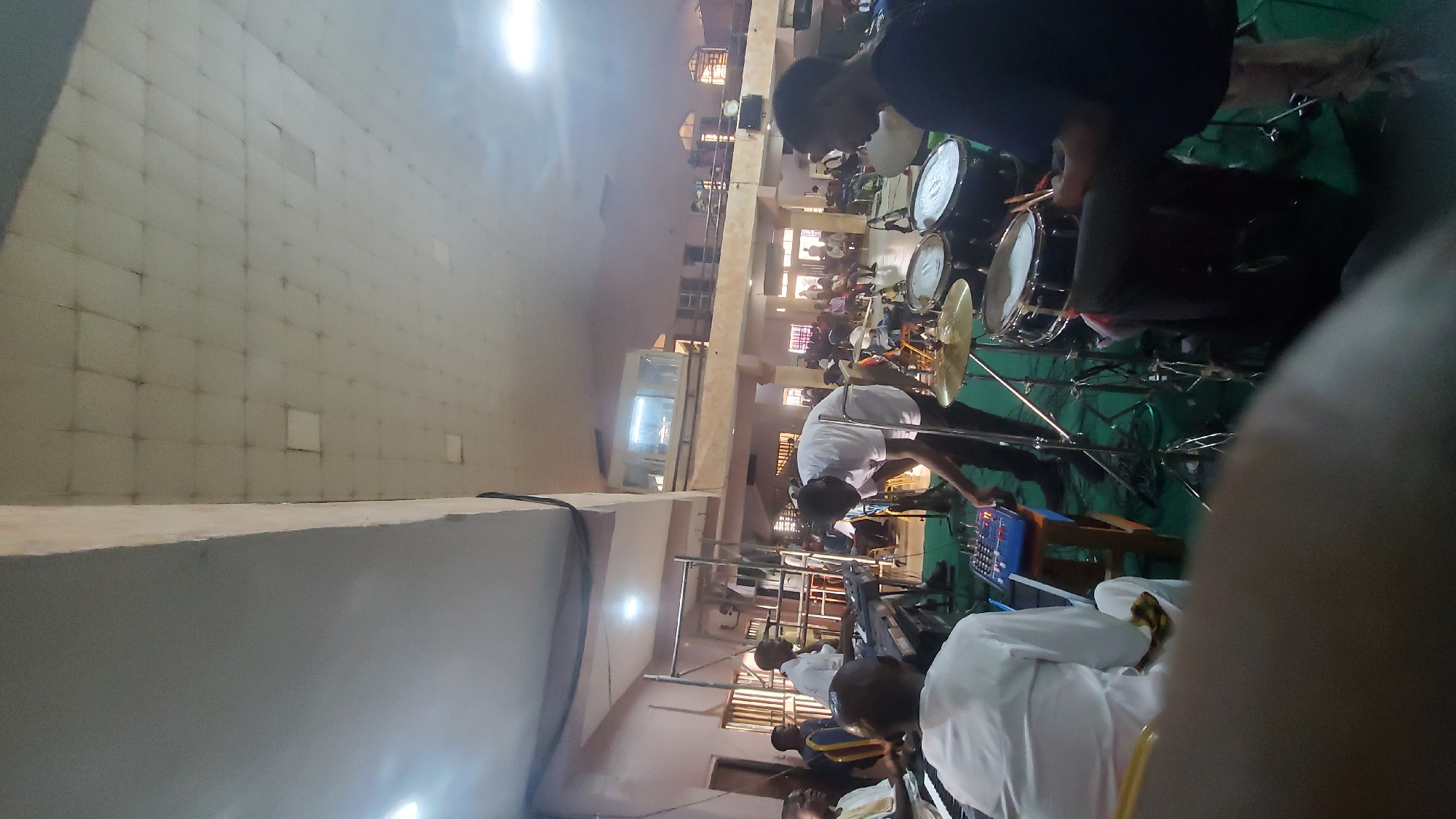 This media file depicts a lively indoor scene, likely a church or event hall, with a band performing. The primary subjects are the band members, mostly young men, dressed in white or dark attire. One individual is actively playing a drum set, with drumsticks in hand. Other band members are positioned behind musical instruments, including keyboards and what appears to be a mixing board. The band is set up on a green surface, possibly a carpet or stage covering.
In the background, a diverse audience is seated, observing the performance. The setting appears to be a large, open space with high ceilings and numerous windows, allowing natural light to filter in, suggesting it is daytime. The architecture features prominent pillars and a multi-level structure, hinting at a substantial building. The overall atmosphere seems energetic and engaging, with the band's performance capturing the audience's attention.
Notable details include the sun's glare visible in some parts of the image, indicating a bright day. The location context of Jalingo, Nigeria, suggests a West African setting. While no explicit text is visible, the scene strongly implies a religious service or a community event. The focus is on the musicians and their instruments, highlighting the dynamic nature of the performance.