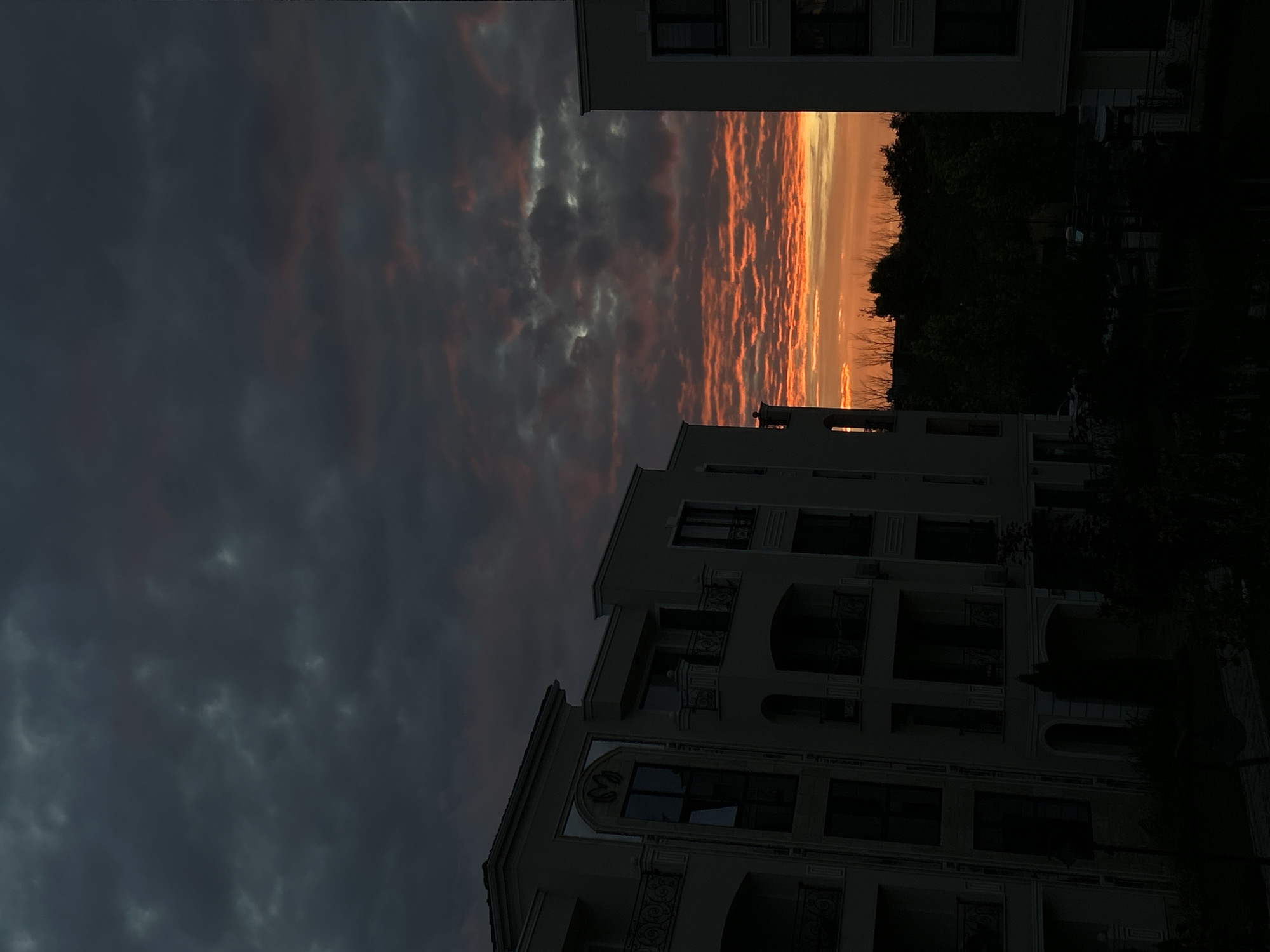 The image depicts a dramatic sunset over a residential area in Plovdiv, Bulgaria. Two modern apartment buildings, constructed with light-colored facades and multiple balconies, flank the view. The sky is filled with dark, ominous clouds that are dramatically illuminated from below by the fiery orange and pink hues of the setting sun. The horizon is dotted with the silhouettes of trees, suggesting a park or natural area between the buildings. The lower portion of the image is in shadow, with only faint outlines of landscaping and possibly parked vehicles visible. The overall impression is one of urban architecture juxtaposed with a powerful natural phenomenon.