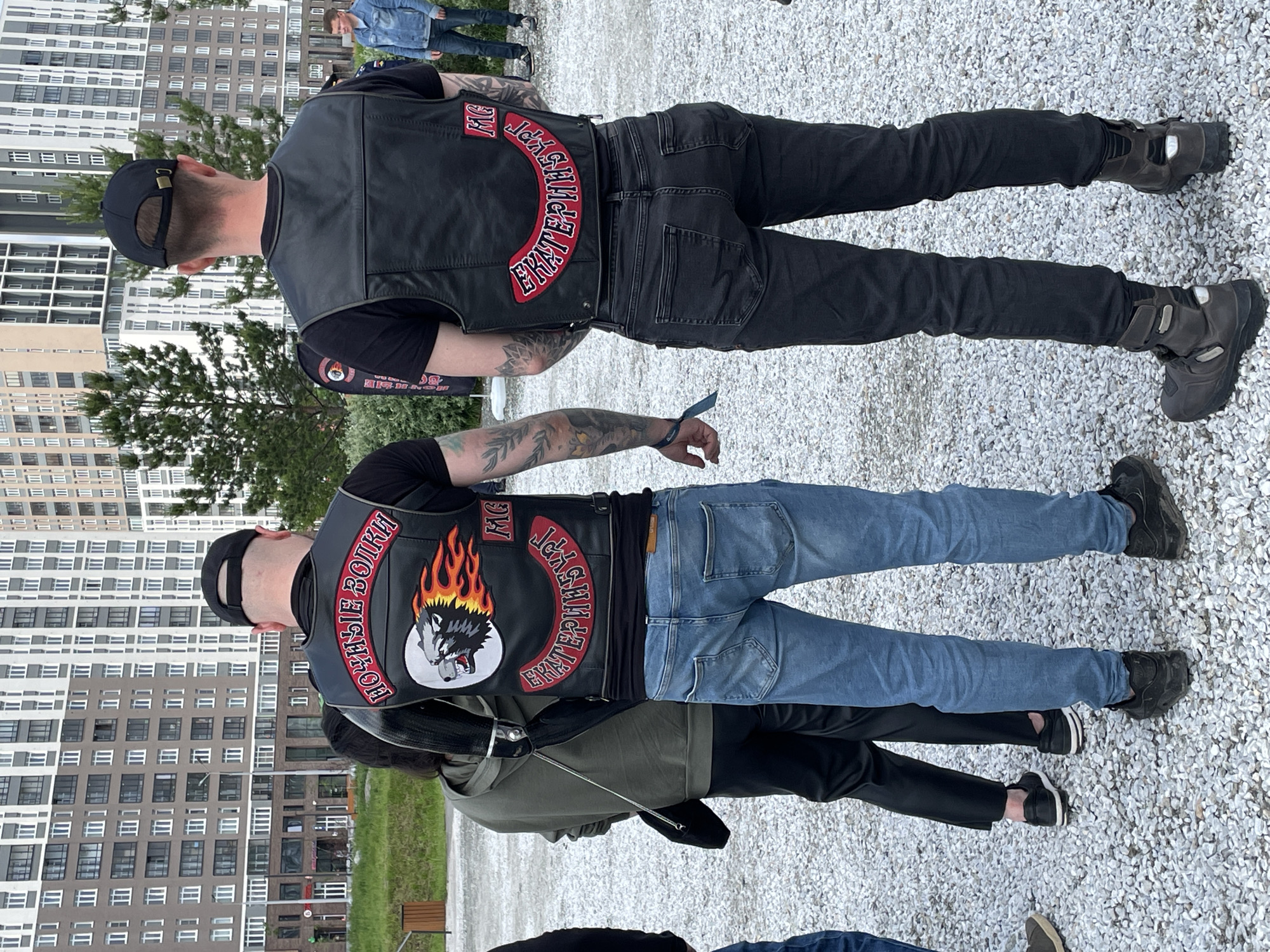 The image captures an outdoor scene in the city of Yekaterinburg, Russia, featuring two men in motorcycle vests viewed from behind on a gravel surface.
The man on the left wears a black baseball cap, a black leather vest, a black short-sleeved t-shirt, light blue denim jeans, and dark sneakers with white soles. His vest features a top rocker patch stating "НОЧНЫЕ ВОЛКИ" (Night Wolves) in red and white, a large central patch depicting a snarling gray wolf head with red and orange flames against a white circle, and a bottom rocker patch stating "ЕКАТЕРИНБУРГ" (Yekaterinburg) in red and white. A small "MC" patch is positioned above the "ЕКАТЕРИНБУРГ" patch. Tattoos are visible on both forearms; the left arm displays a leafy pattern, while the right arm has a more abstract design. A blue wristband is present on his left wrist.
The man on the right, positioned slightly further back, also wears a black baseball cap, a plain black leather vest, a black short-sleeved t-shirt, dark gray/black slim-fit jeans, and dark brown or black motorcycle boots with visible buckles and straps. His vest has a small "MC" patch in red and white positioned on the upper right side and a bottom rocker patch stating "ЕКАТЕРИНБУРГ" (Yekaterinburg) in red and white. Tattoos are visible on both forearms with dark, patterned designs.
Between these two men, slightly to the left, a woman is partially visible from behind. She wears a dark olive-green hooded sweatshirt, black pants, and dark sneakers with white soles. A small black purse with a silver chain strap hangs from her left shoulder.
The ground consists of light-colored gravel. In the background, multiple tall, modern residential buildings with varied facades (light and dark panels) are visible. Sparse green grass and several small evergreen trees are situated between the gravel area and the buildings. A dark brown waste receptacle is visible on the far left. A street light pole is present in the mid-ground, and a blurred banner with red and black elements is partially visible behind the right man. On the far right, a man in a light blue denim jacket and blue jeans is visible in the distance.
