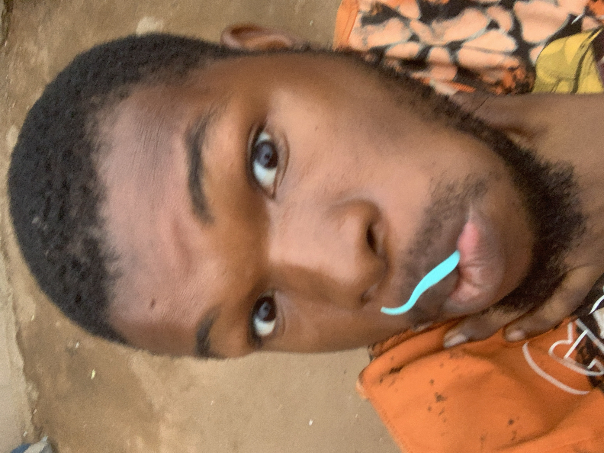 A close-up portrait depicts a person with dark skin and short, dark hair. They have a beard and are looking directly at the camera. A light blue, curved object is positioned near their mouth. The person is wearing an orange garment with a white design. The background consists of an earth-toned surface and a patterned fabric or garment with orange, brown, and yellow hues.
This image does not provide enough information to determine a precise location in Onitsha, Nigeria.