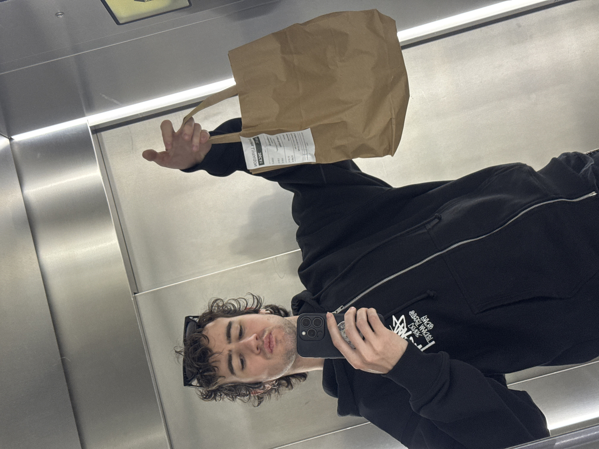 A young man with curly hair, wearing a black zip-up hoodie and sunglasses perched on his head, takes a selfie in a mirrored environment, likely an elevator. He holds an iPhone in his right hand and a brown paper bag with a white label in his left hand. The elevator's interior appears to be brushed metal. The photo was taken in Paris, France.