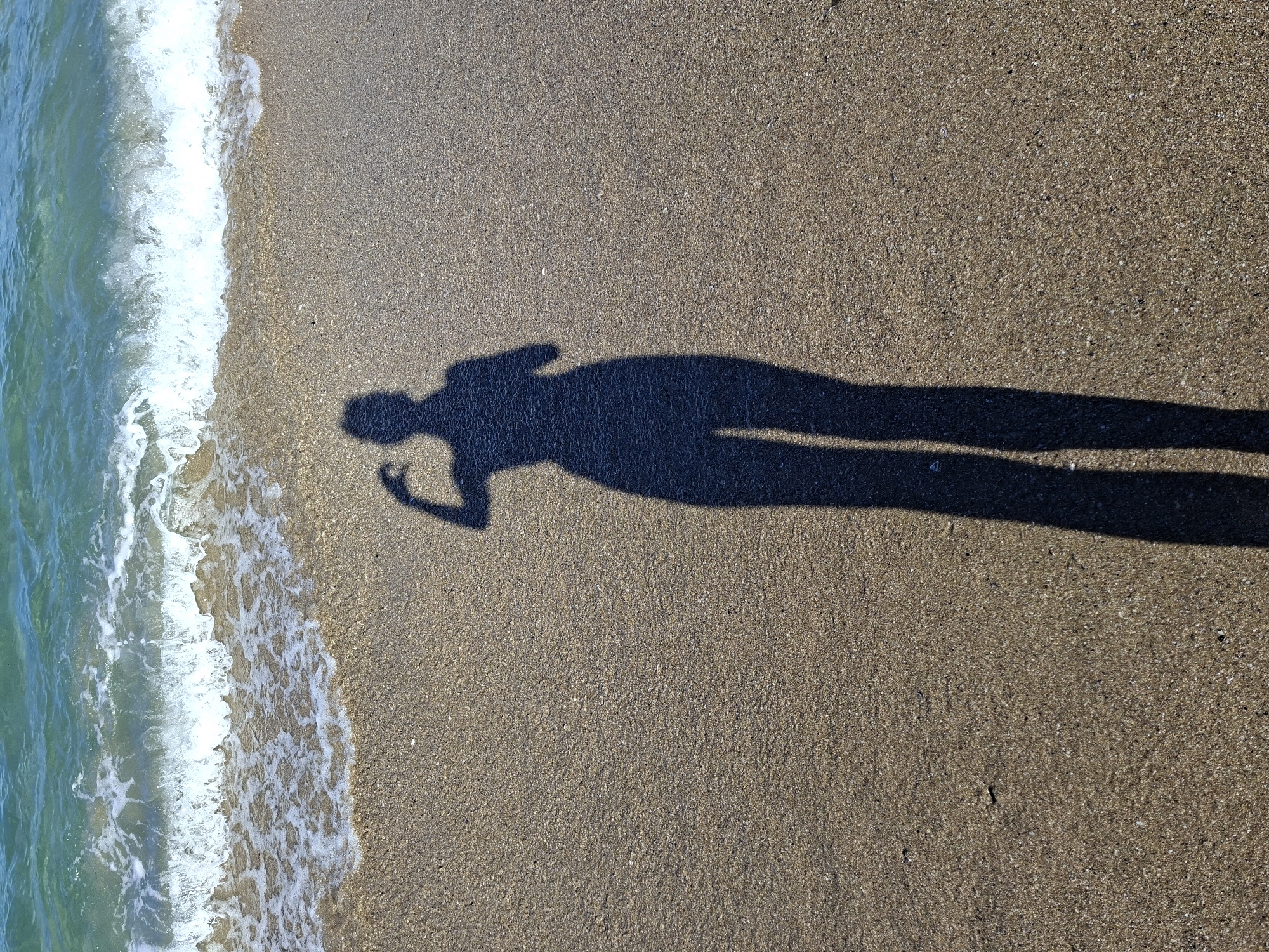 A shadow of a person, possibly female, is cast on a sandy beach. The shadow is flexing its left bicep. The person is standing near the water's edge, with a wave gently lapping at the shore. The water is a clear turquoise color and appears calm. The sand is a light brown and looks damp in some areas. The scene is illuminated by bright sunlight, creating a strong shadow. The location is Unknown, Unknown.