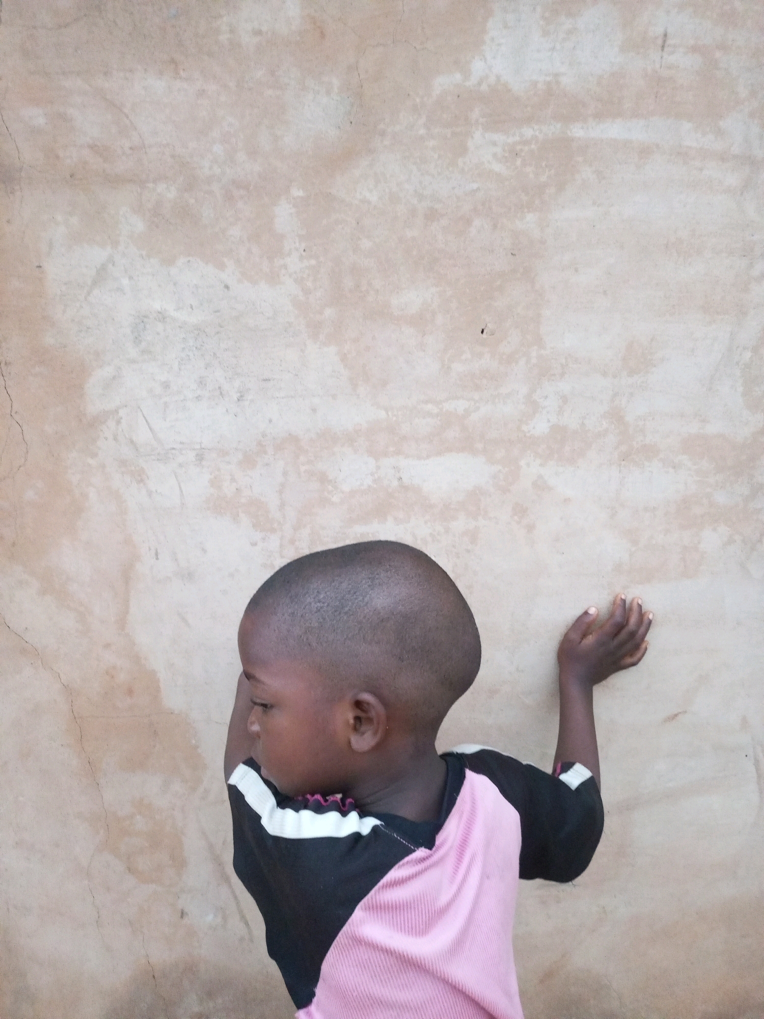 A child, viewed from behind and slightly to the side, leans against a textured, light brown wall. The child has short, dark hair and is wearing a black t-shirt with white stripes across the shoulders and upper chest, and a pink, ribbed material on the lower part of the shirt. The child's right arm is extended, with the hand resting on the wall. The child's left arm is bent at the elbow, with the hand near their side. The wall appears to be made of rough plaster and has some visible cracks and variations in color. The scene is lit by natural light, casting subtle shadows. The image is cropped to focus on the child and the wall. This image was taken in Tempure, Nigeria.