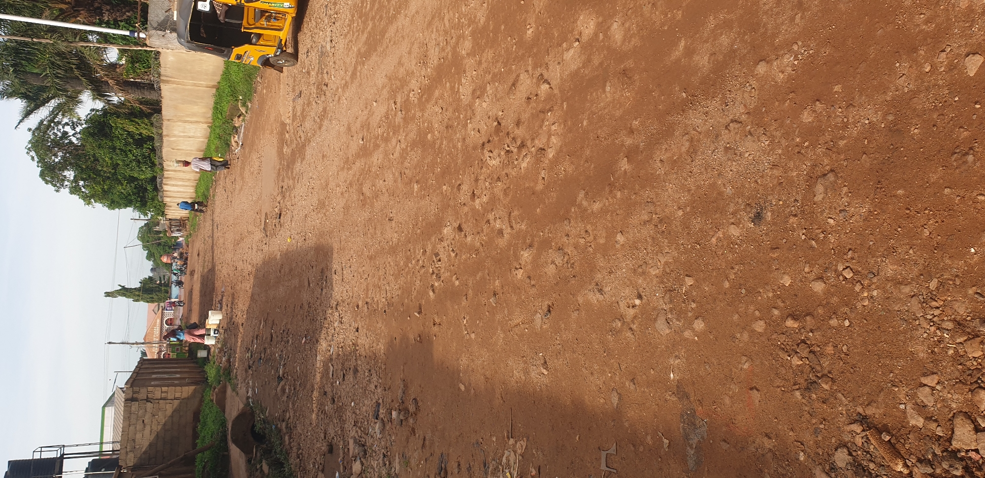 The image depicts a wide, unpaved reddish-brown dirt road in Sabon Tasha, Nigeria, under clear daylight conditions. The road surface is uneven, characterized by visible rocks, gravel, and minor depressions. Shadows cast from objects on the left suggest a high sun angle.
On the left side of the road, a low concrete block wall extends into the mid-ground. Behind this wall, a metal frame supports two black water tanks. Adjacent to the wall, a person wearing a light blue top and dark pants is attending to several white plastic buckets arranged on the ground. Further down the road on the left, additional buildings with varied rooflines, including one with a reddish tiled roof, are visible, interspersed with green vegetation. Utility poles with power lines run along this side of the street. In the distant background on the left, a gathering of people and several motorcycles indicates further activity.
On the right side of the road, a tall, light-colored concrete wall topped with barbed wire runs parallel to the path. Green grass and foliage grow at its base. A person is walking away from the foreground, balancing a large, clear water container on their head and holding a yellow object in their hand. Further ahead, another person is seen riding a motorcycle. Partially visible in the extreme foreground on the right is the front section of a yellow and black three-wheeled auto-rickshaw. Behind the wall on the right, dense green trees, including palm trees, are present.
The overall scene captures daily life and infrastructure in a residential or semi-commercial area, with a focus on local transportation and water collection activities.