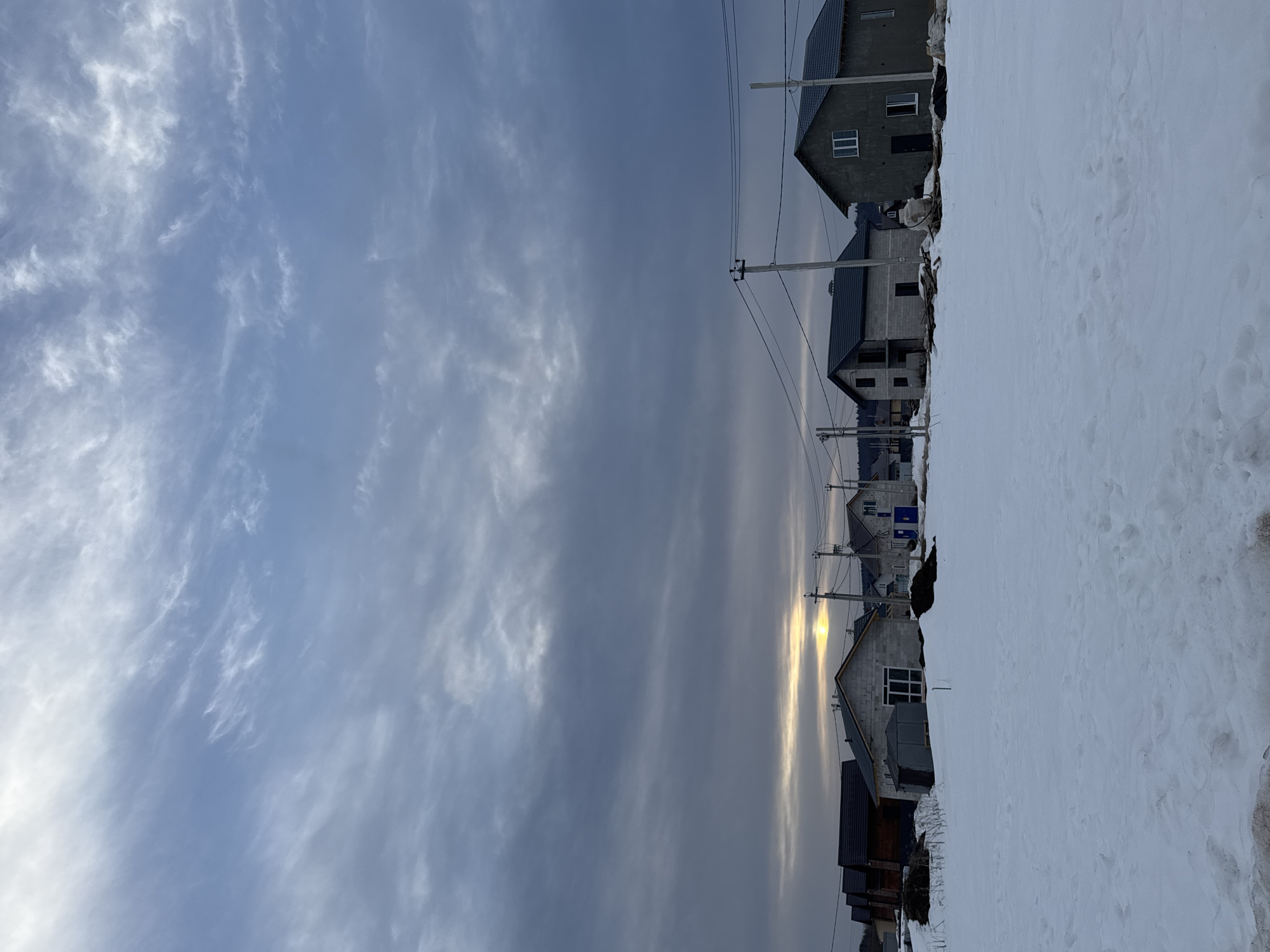 The image depicts a winter scene in Gornyi (Dvurechenskoe S/P), Russia, characterized by an expansive, snow-covered foreground with minimal visible tracks. The midground features a developing residential street lined with approximately five houses. Most structures are two-story, constructed primarily from light grey concrete blocks, with dark, sloped roofs, suggesting ongoing construction or recent completion.
From left to right, the visible buildings include a darker-colored house with a dark roof, followed by a lighter grey concrete block house with a visible window opening and a small, dark utility shed positioned to its left. Further right, another grey concrete block house with a dark roof is partially obscured by utility poles. The furthest visible complete house on the right is finished with a dark grey render, featuring white-framed windows and a dark entrance door. Several utility poles with overhead power lines run horizontally across the midground, between and in front of the houses. Piles of disturbed earth or debris are visible near some of the structures.
The background shows a distant line of trees or forest along the horizon. The sky is largely overcast with dense, varied grey and blue cloud formations. A bright, localized yellow-orange glow, indicating either a low sun at sunrise or sunset, is visible just above the horizon, partially obscured by the cloud cover. No individuals, actions, or direct interactions are visible within the scene.