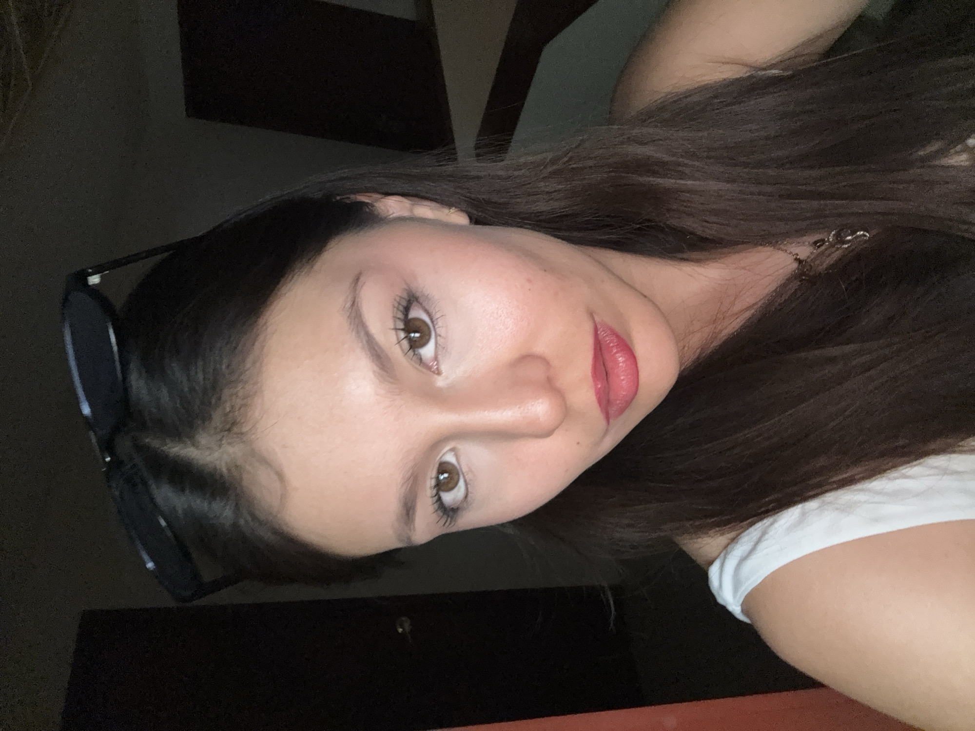 A close-up, low-angle, indoor photograph depicts a young woman with fair skin and dark brown hair. Her hair is long and loose, partially obscuring her right shoulder, which is covered by a white garment. She has brown eyes, full lips with reddish lipstick, and sunglasses partially visible on the top of her head. The background is dark, featuring a door and a light switch or handle. The setting is assumed to be an indoor location in Heroica Puebla de Zaragoza, Mexico, based solely on user context and not discernible from the image itself.