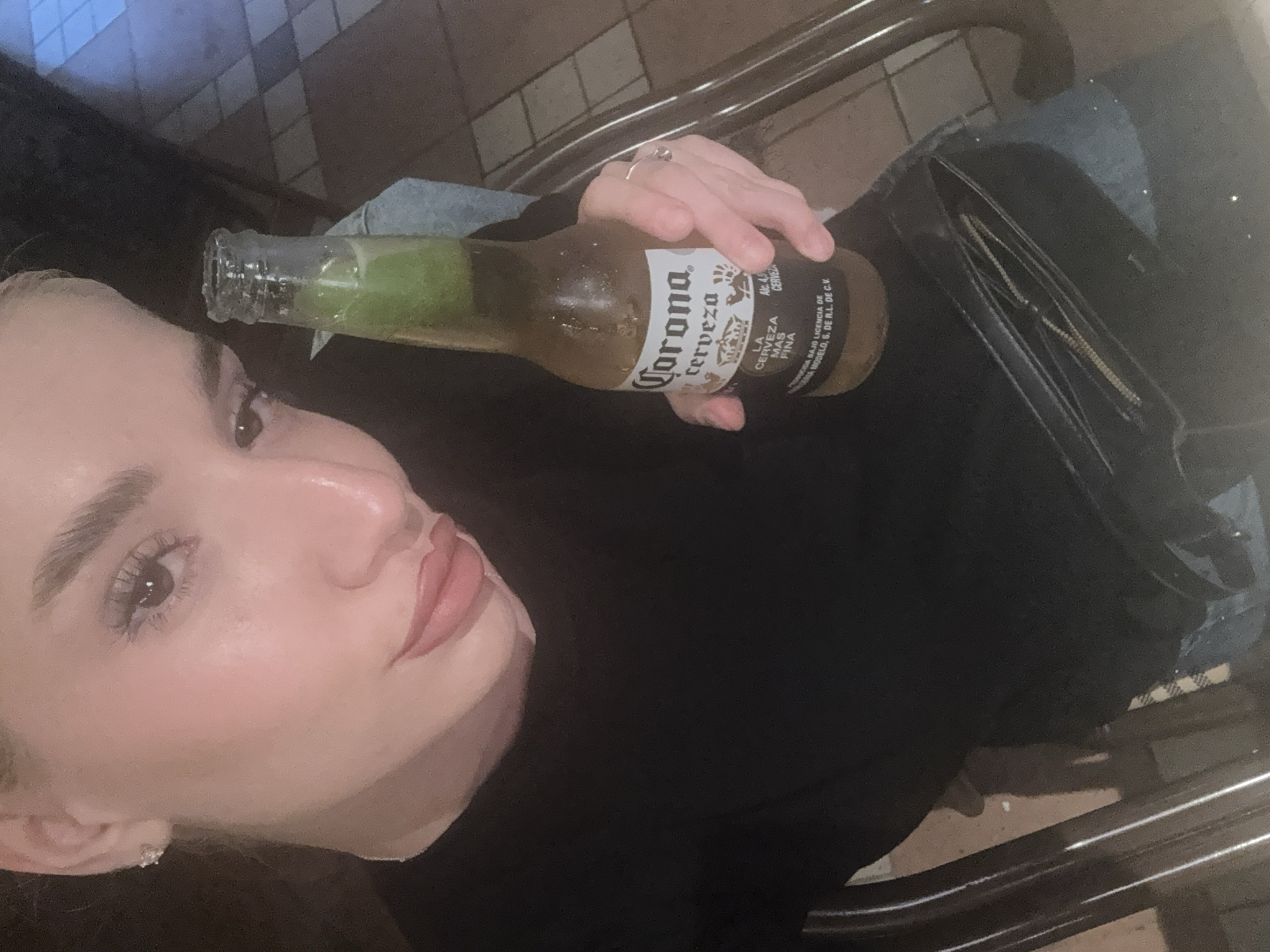 The image captures a close-up, high-angle selfie of a woman holding a bottle of Corona beer, likely taken in a dimly lit indoor public space in Sofia, Bulgaria.
The primary subject is a woman, seen from the mid-chest up, looking towards the camera. She has light skin, dark eyebrows, and is wearing makeup including mascara and a light-colored lip product. She wears a ring on her right ring finger and a small stud earring. Her attire includes a dark-colored top and light-washed denim jeans.
The main object she is holding is a glass bottle of Corona Cerveza, which appears cold as indicated by condensation. A lime wedge is visible inside the bottle's neck. A black purse or bag with a zipper is also visible, resting on her lap or beside her.
The background suggests an urban setting, possibly a bar, restaurant, or transportation interior. Details include a tiled wall and metal rails, implying a casual environment. The lighting is dim, suggesting it is evening or nighttime.
The activity taking place is the woman posing for a self-portrait while holding her drink. Her expression is neutral, common for a selfie.
Visible text on the beer bottle label reads: "Corona cerveza LA CERVEZA MAS FINA Alc. 4,5 CERVEZA", followed by smaller text "HECHA EN MEXICO BAJO LICENCIA DE GRUPO MODELO, S. DE R.L. DE C.V.".