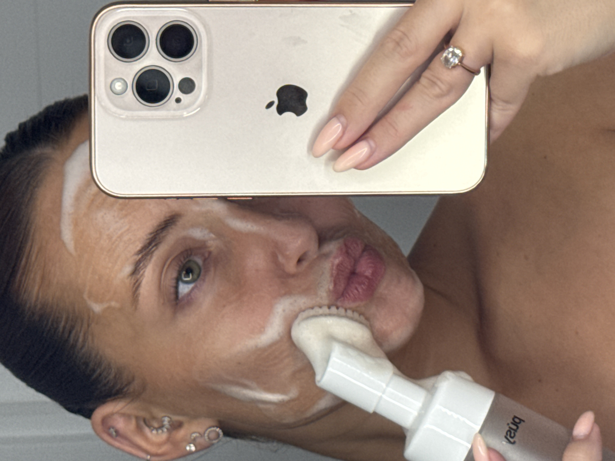 The image shows a close-up of a woman's face as she cleanses her skin. She is holding a white foaming cleanser with a brush applicator in her left hand and using her right hand to hold up a smartphone, likely taking a selfie. The woman has her hair pulled back, and her face is partially covered in white foam. She is wearing multiple earrings in her left ear and a ring on her right ring finger. The background is nondescript, appearing to be an indoor setting. There is no visible indication of the location being Chaikovsky, Russia.